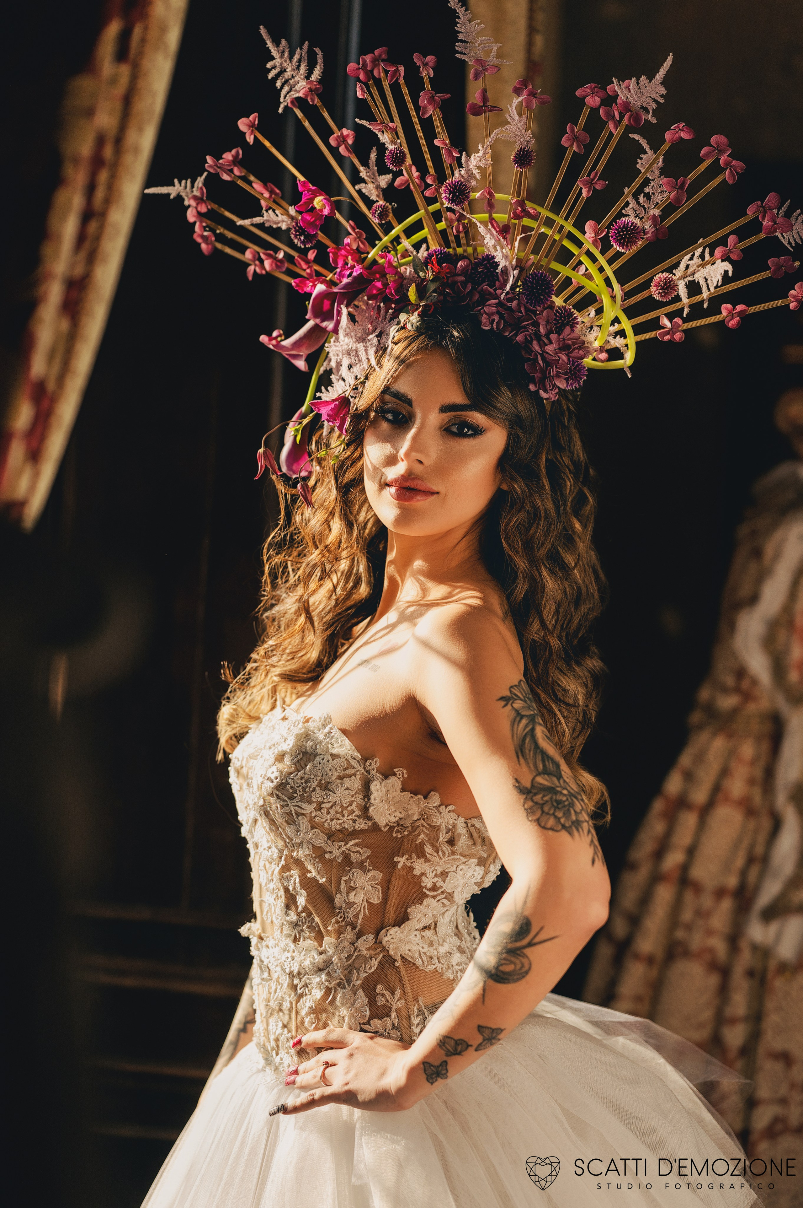 Sposa con acconciatura floreale extra-large in toni rosso e rosa indossa un abito da sposa in pizzo con schiena scoperta durante uno shooting di matrimonio artistico.