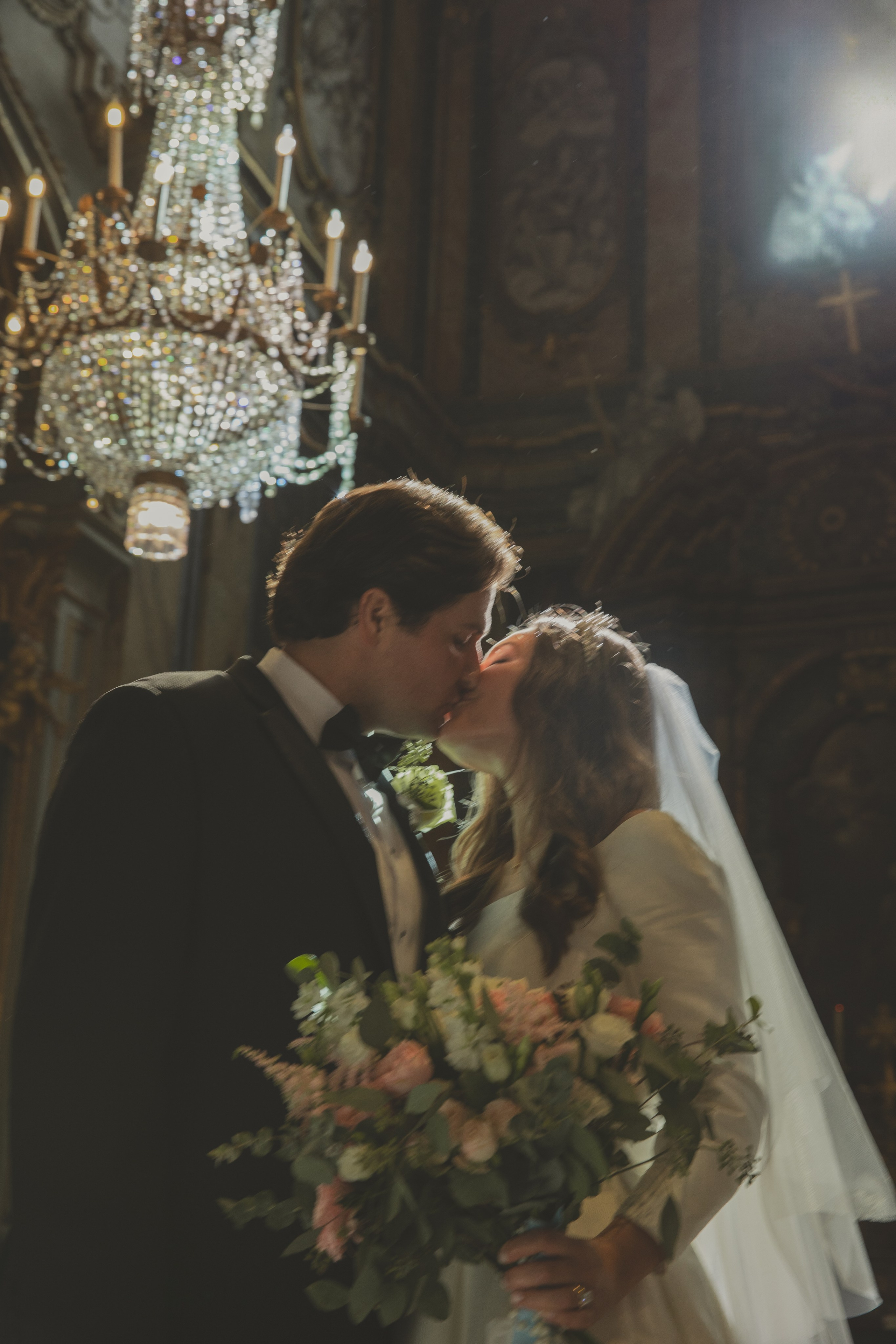 Romantic bride and groom kiss inside a historic Roman church during their elegant destination wedding in Italy. The bride holds a lush bouquet of pastel flowers, bathed in soft golden light under a grand chandelier.