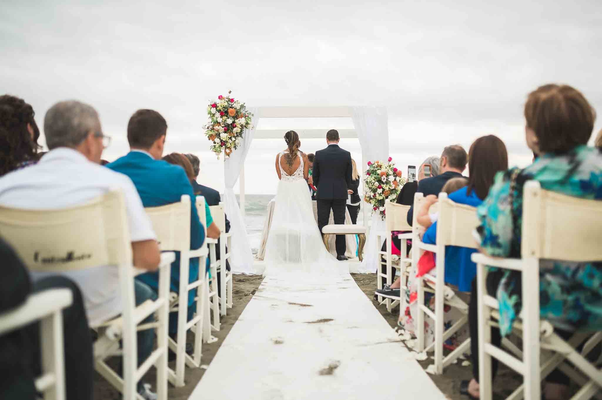 Perspettiva degli invitati durante una cerimonia nuziale all'aperto, con la coppia di sposi di fronte a un arco fiorito sullo sfondo del mare, simbolo di nuovi inizi e promesse condivise.