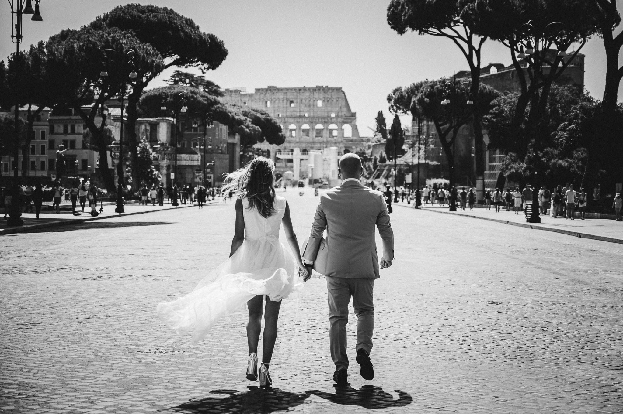 Neosposi mano nella mano avanzano verso il Colosseo in una giornata soleggiata, catturati in una fotografia in bianco e nero dal loro fotografo di matrimonio; i dettagli del vestito da sposa che fluttua al vento e l'eleganza del vestito dello sposo si stagliano contro l'antico sfondo romano.