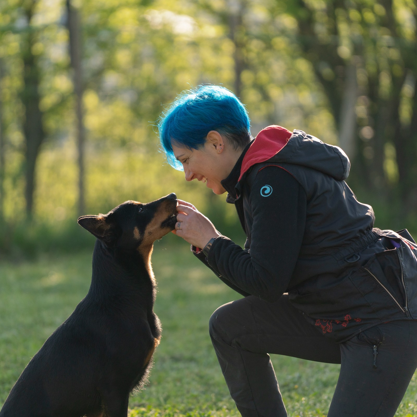 Una addestratrice dai capelli blu è inginocchiata sull'erba mentre interagisce con un cane nero e marrone. Sembra offrire o dare qualcosa al cane, che sta in piedi e guarda verso di lei con attenzione. Lo sfondo è composto da alberi e vegetazione, dando alla scena un'atmosfera naturale e rilassante. Il momento cattura un legame speciale tra l'addestratrice e il cane, suggerendo un momento di insegnamento o affetto.