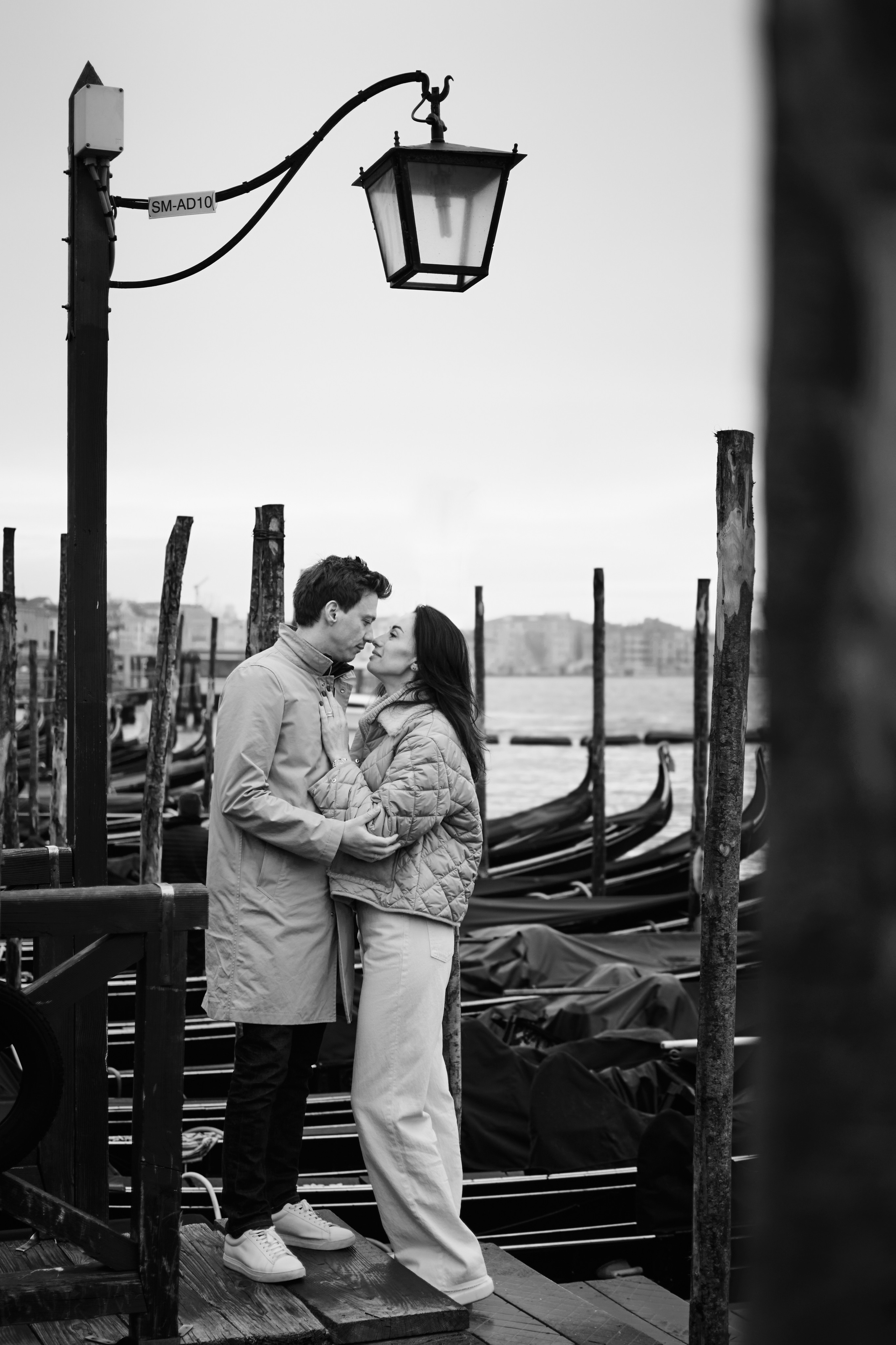 A couple in love stands hand in hand on San Marco Square, Venice, with the cloudy sky in the background. The grand architecture of Venetian canal  rises up behind them. The couple is lost in each other's eyes, oblivious to the bustling crowds around them. The grey clouds overhead create a moody and romantic atmosphere, as if the heavens themselves are blessing their love.
