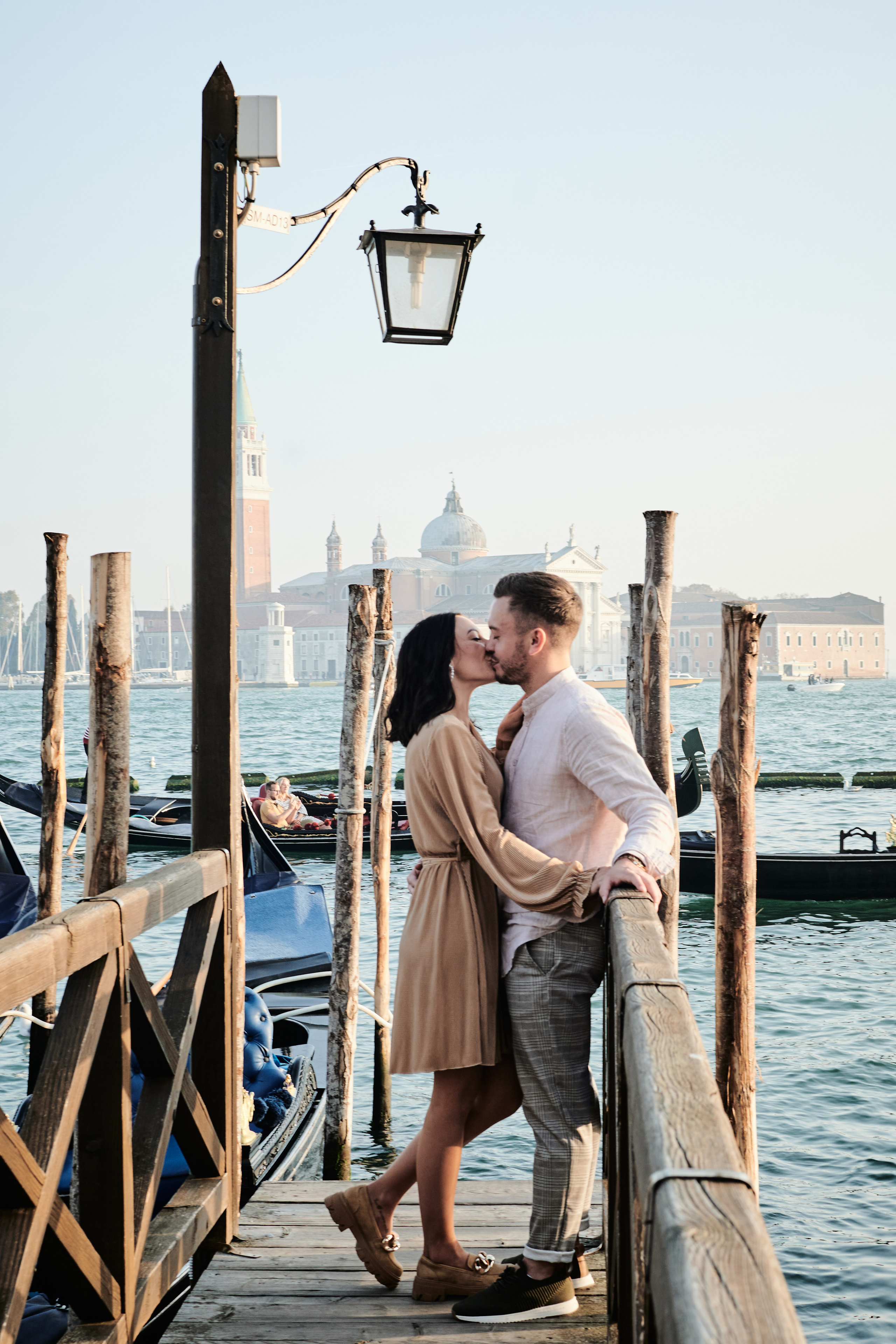 In this beautiful portrait photo, a couple stands hand-in-hand, gazing out at the breathtaking panorama of the lagoon and San Marco Square. The intricate details of the surrounding architecture and the warm, golden light of the setting sun create an atmosphere of beauty, love, and passion