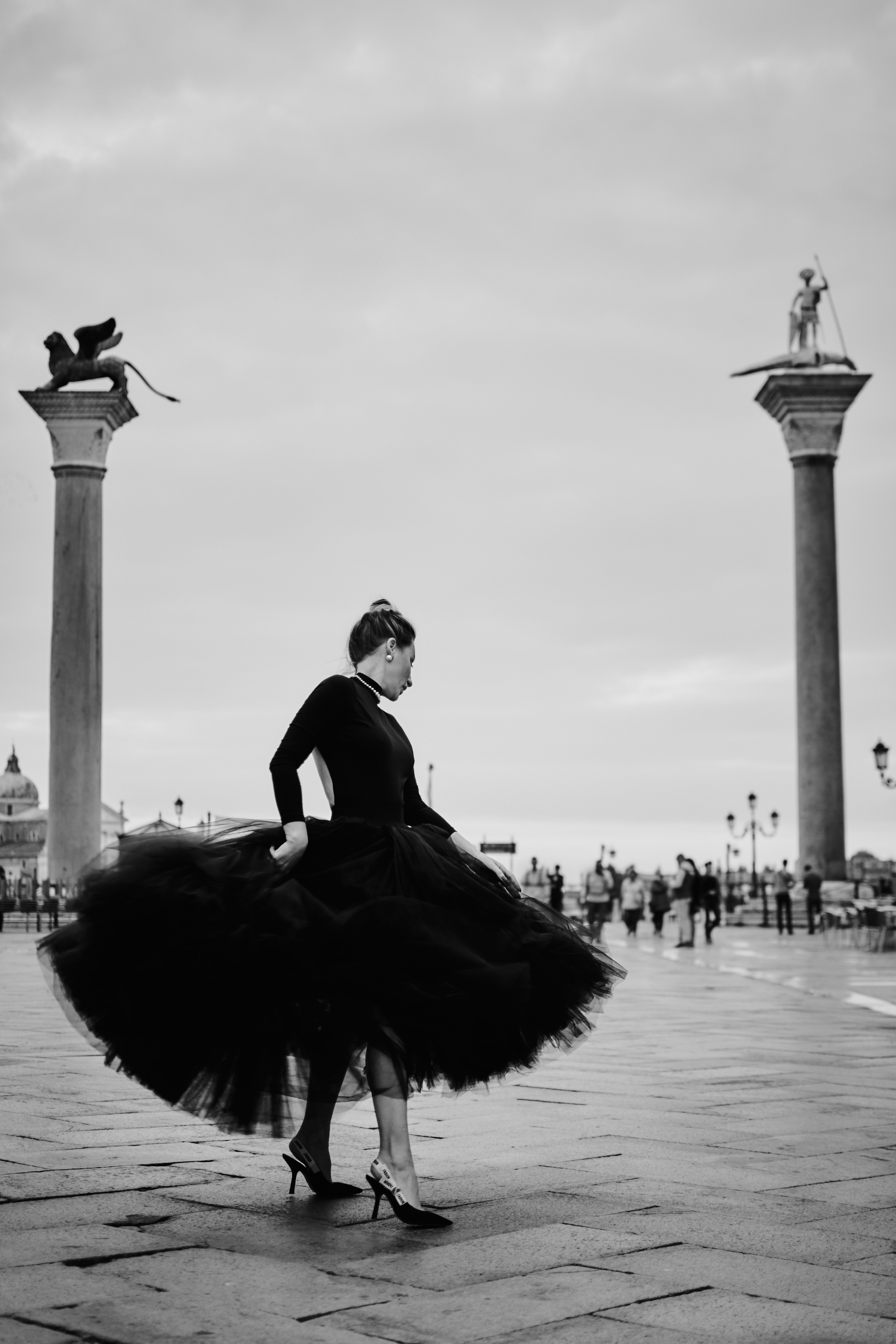 A young woman in a black dress stands gracefully in front of the arches of San Marco Square in Venice. The intricate details of the architecture frame her figure in a timeless elegance, while the soft light of the setting sun illuminates her features. Her dark hair cascades down her shoulders, and her eyes are fixed on some distant point, lost in thought. grand Columns of San Marco and San Todaro on the iconic San Marco Square in Venice.The scene captures the romantic and captivating essence of Venice's charm, with the woman's presence adding a touch of mystery and allure to the already enchanting backdrop.