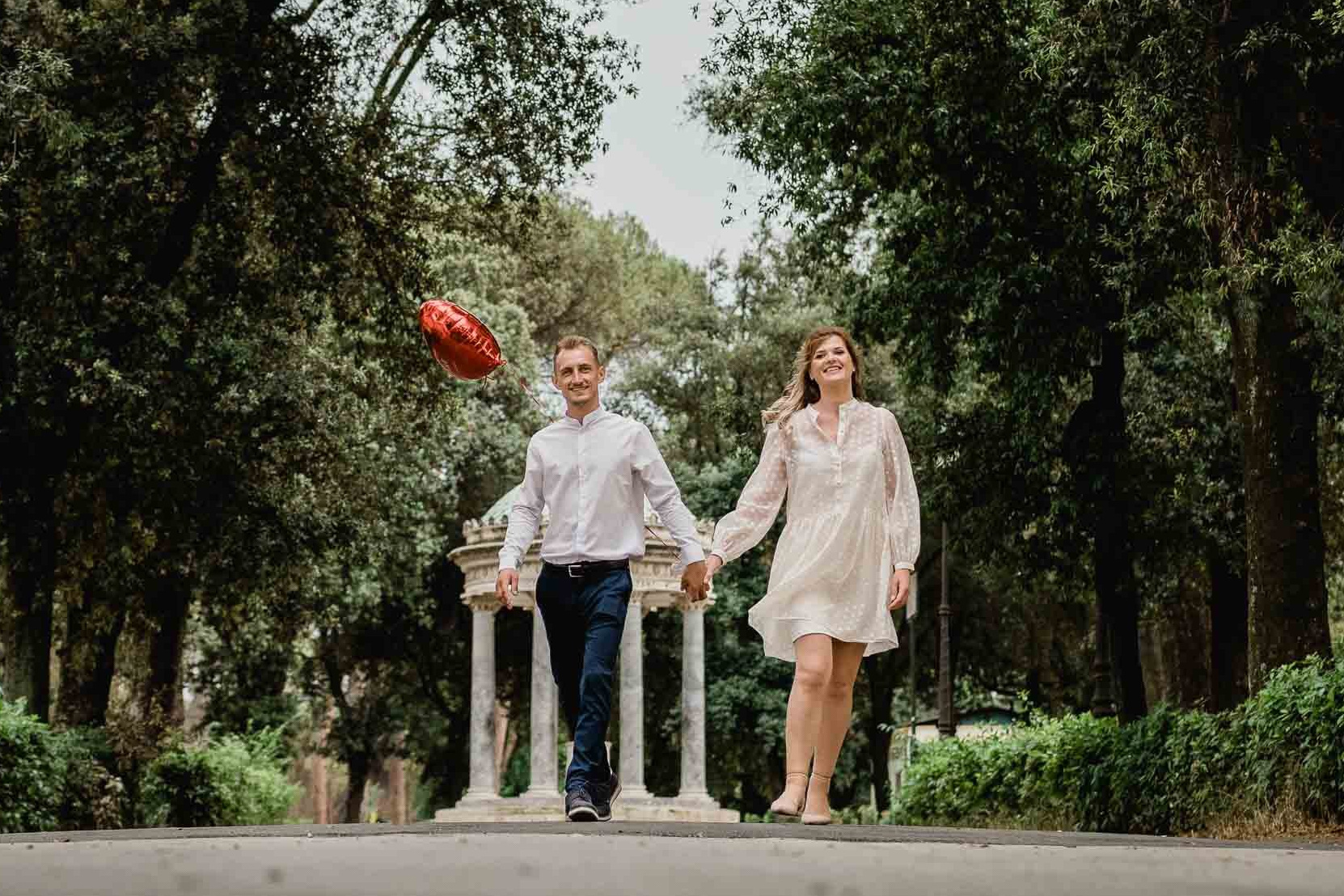 Neosposi mano nella mano avanzano verso il Colosseo in una giornata soleggiata, catturati in una fotografia in bianco e nero dal loro fotografo di matrimonio; i dettagli del vestito da sposa che fluttua al vento e l'eleganza del vestito dello sposo si stagliano contro l'antico sfondo romano.