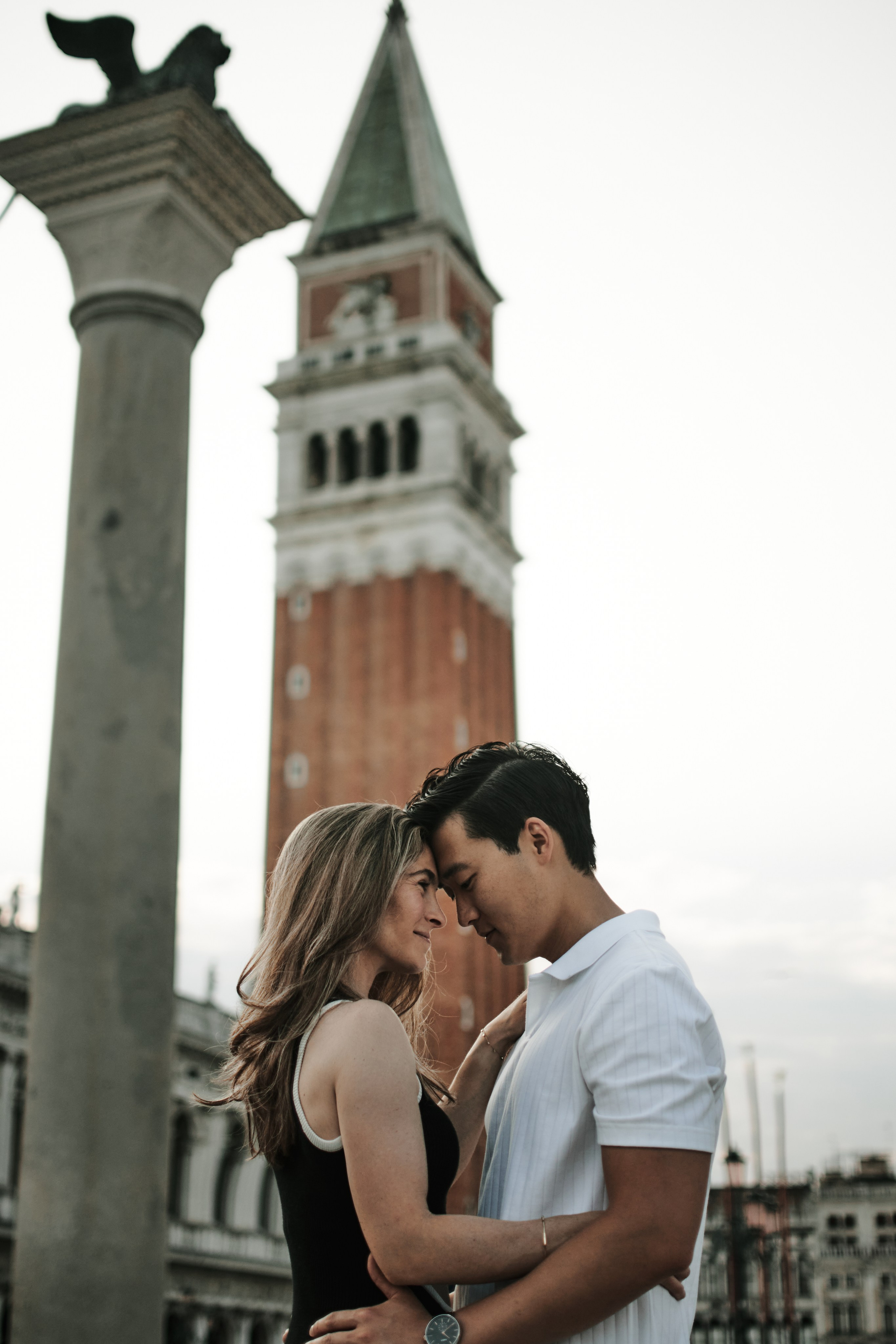 A romantic portrait captures a couple in the midst of a love story shoot on San Marco Square. They stand close together, gazing out at the iconic Bridge of Sighs in the distance. The photographer has expertly framed the shot to showcase both the couple's love and the beauty of Venice's architecture