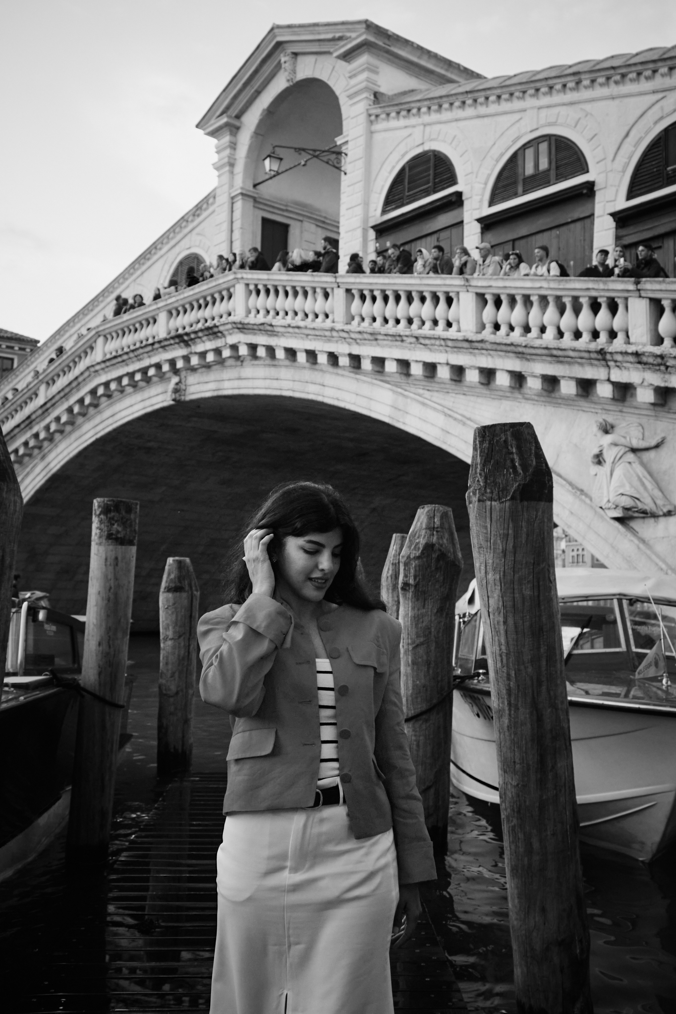 A young woman stands near Rialto Bridge in Venice, Italy. She wears a flowing, floral dress that catches the gentle breeze blowing in from the sea. Her hair is styled in loose waves and she holds a delicate parasol to shield her from the sun. In the background, the iconic St. Mark's Basilica stands tall, its intricate architecture and golden mosaics glistening in the sunlight. The bustling square is filled with people and the sounds of music and laughter, creating a lively and romantic atmosphere.