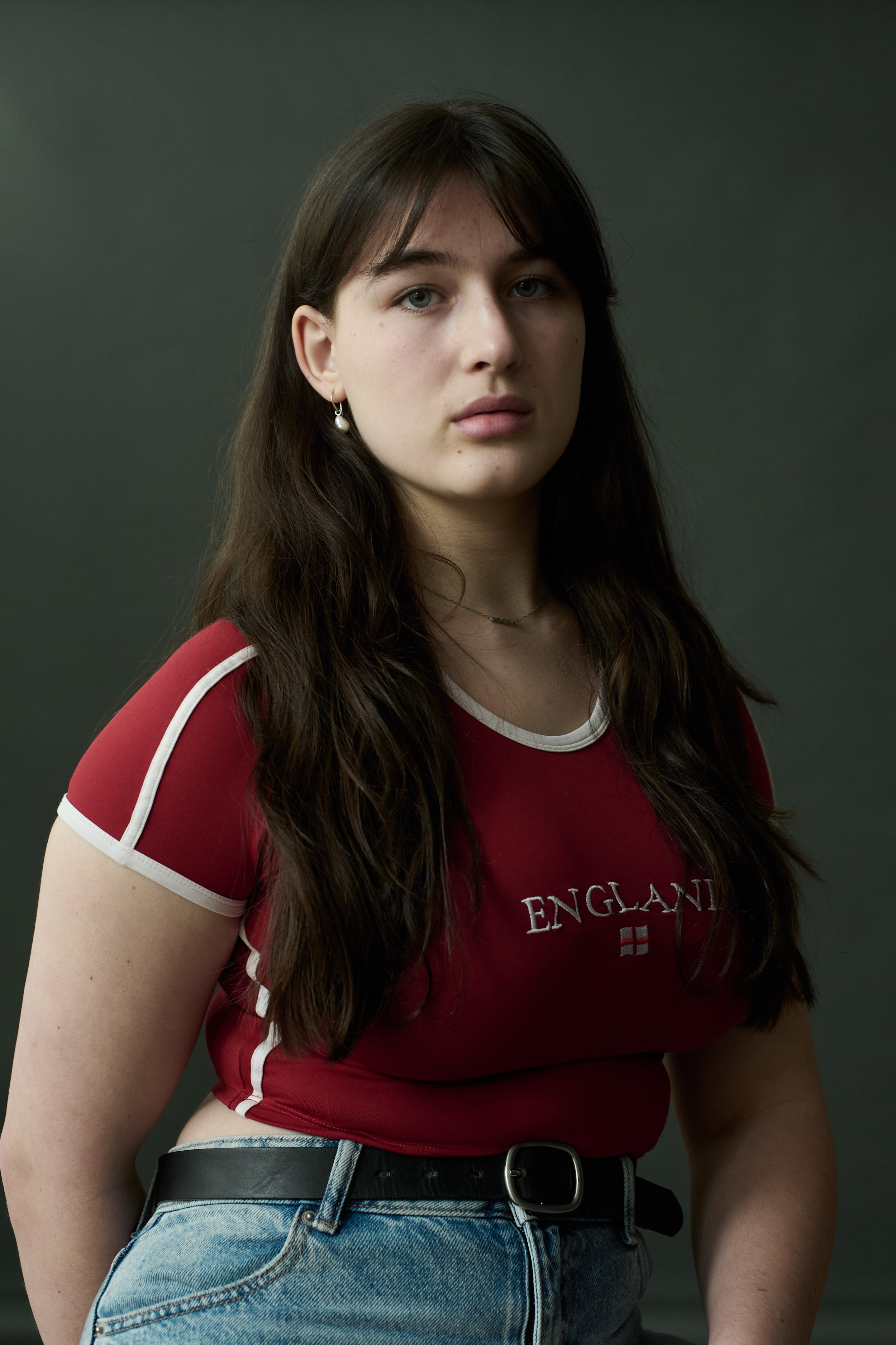 Photographer in Brussels and Amsterdam, Victoria Ushkanova.Portrait of a young woman with a pearl earring sitting on a stool, wearing a red “England” shirt and jeans 