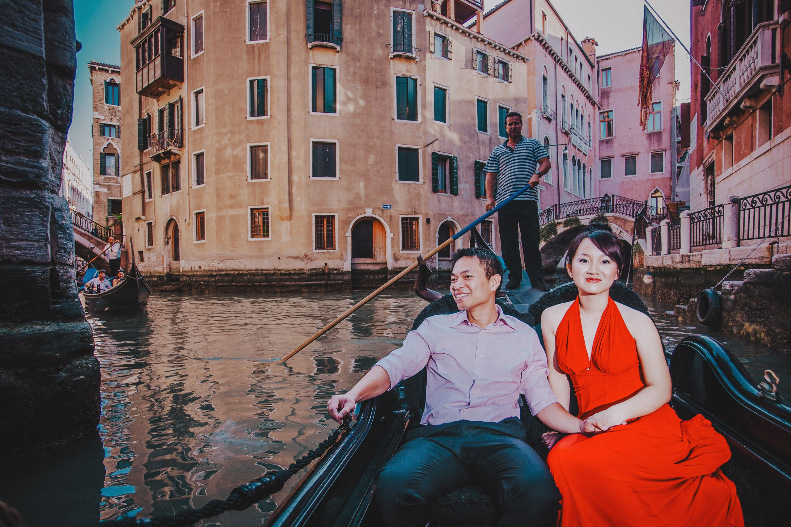 A Thai woman wearing a bold red dress holds hands with her fiancee as they explore the canals of Venice in a gondola during their Love Story adventure in Italy.