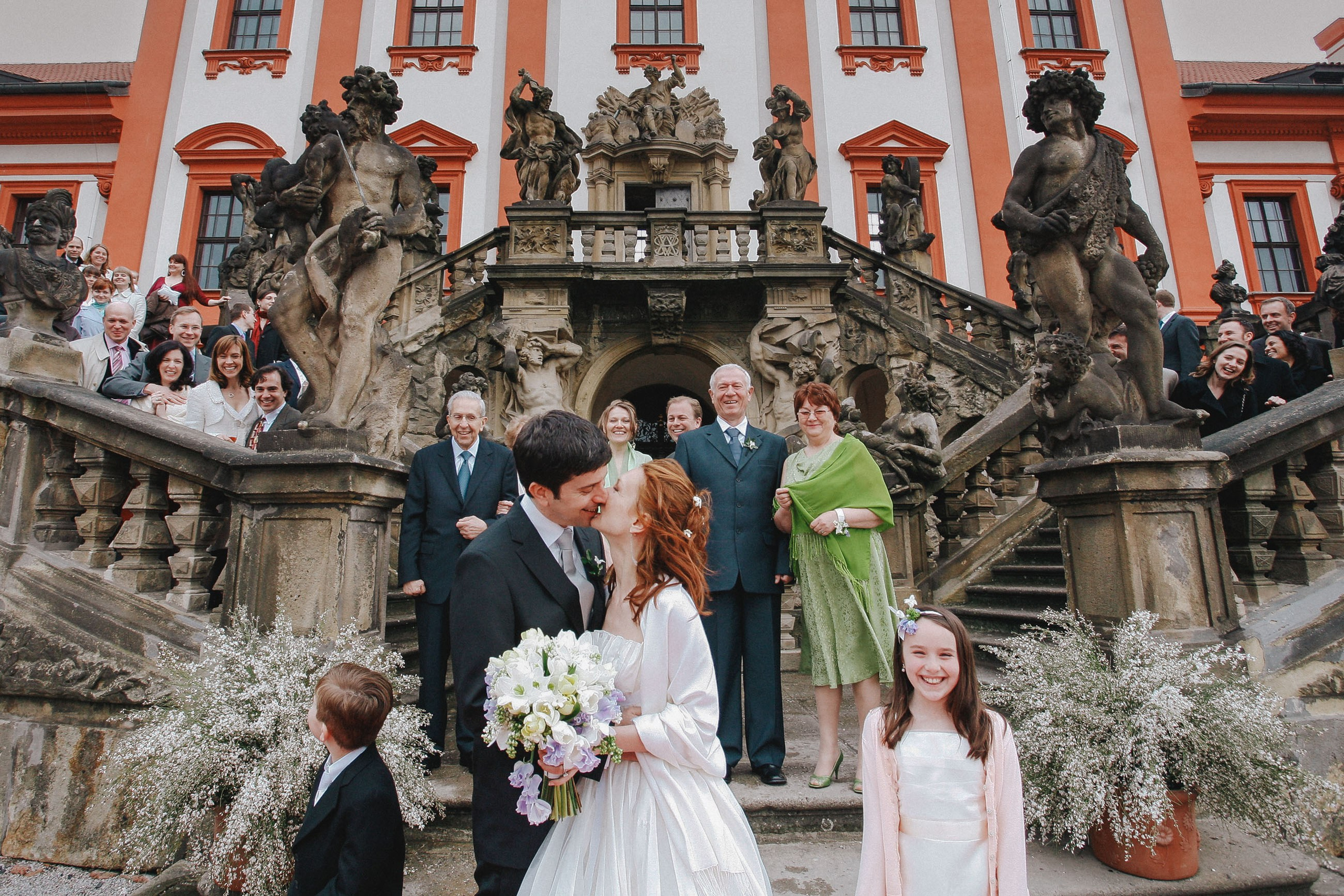 American groom kissing Russian bride on Toja Castle steps, surrounded by celebrating family and friends.