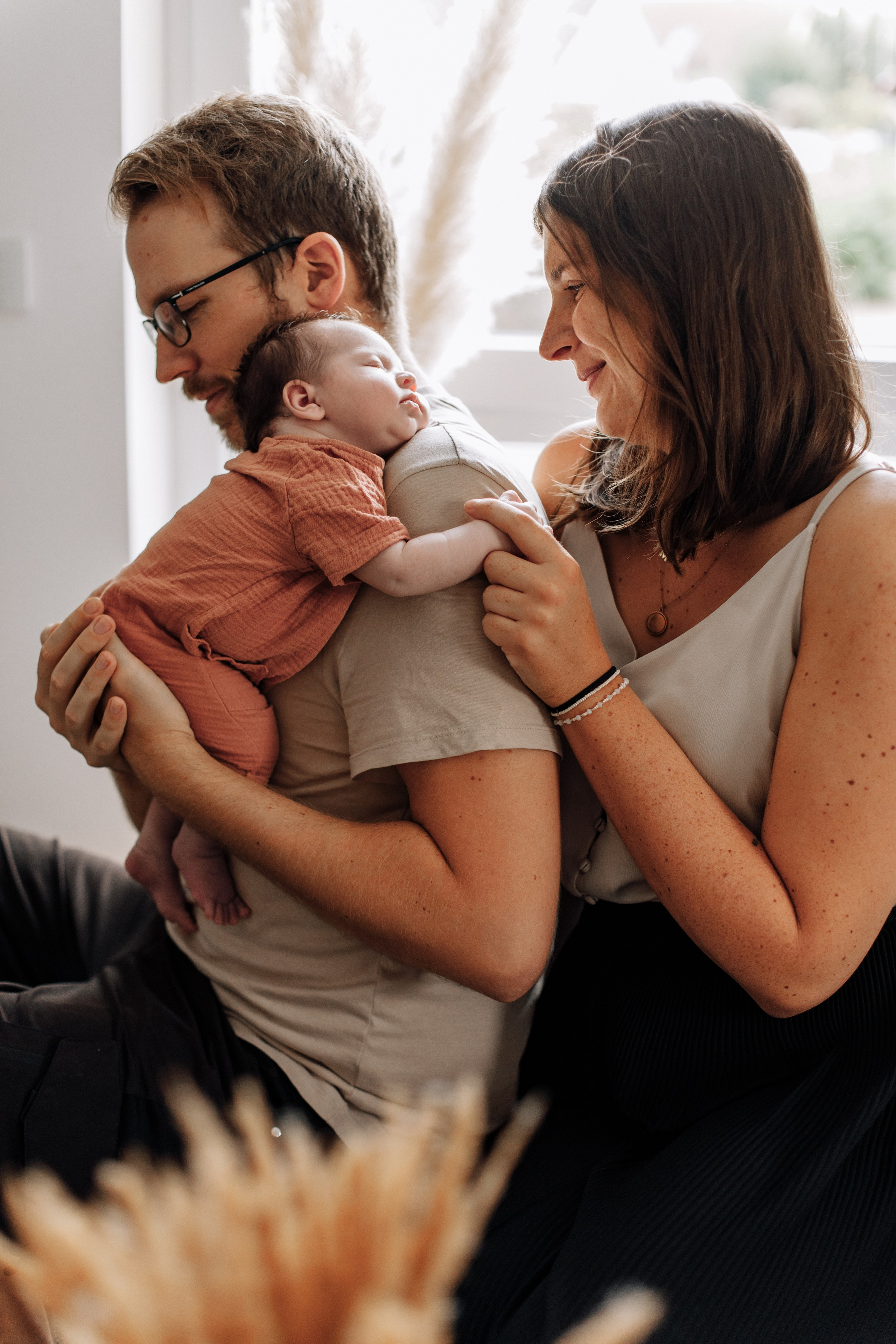 Mädchen schläft auf der Schulter des Vaters, während die Mutter ihre Hand hält. Sie sitzen gemeinsam am Fenster während eines Familienshootings zu Hause. 