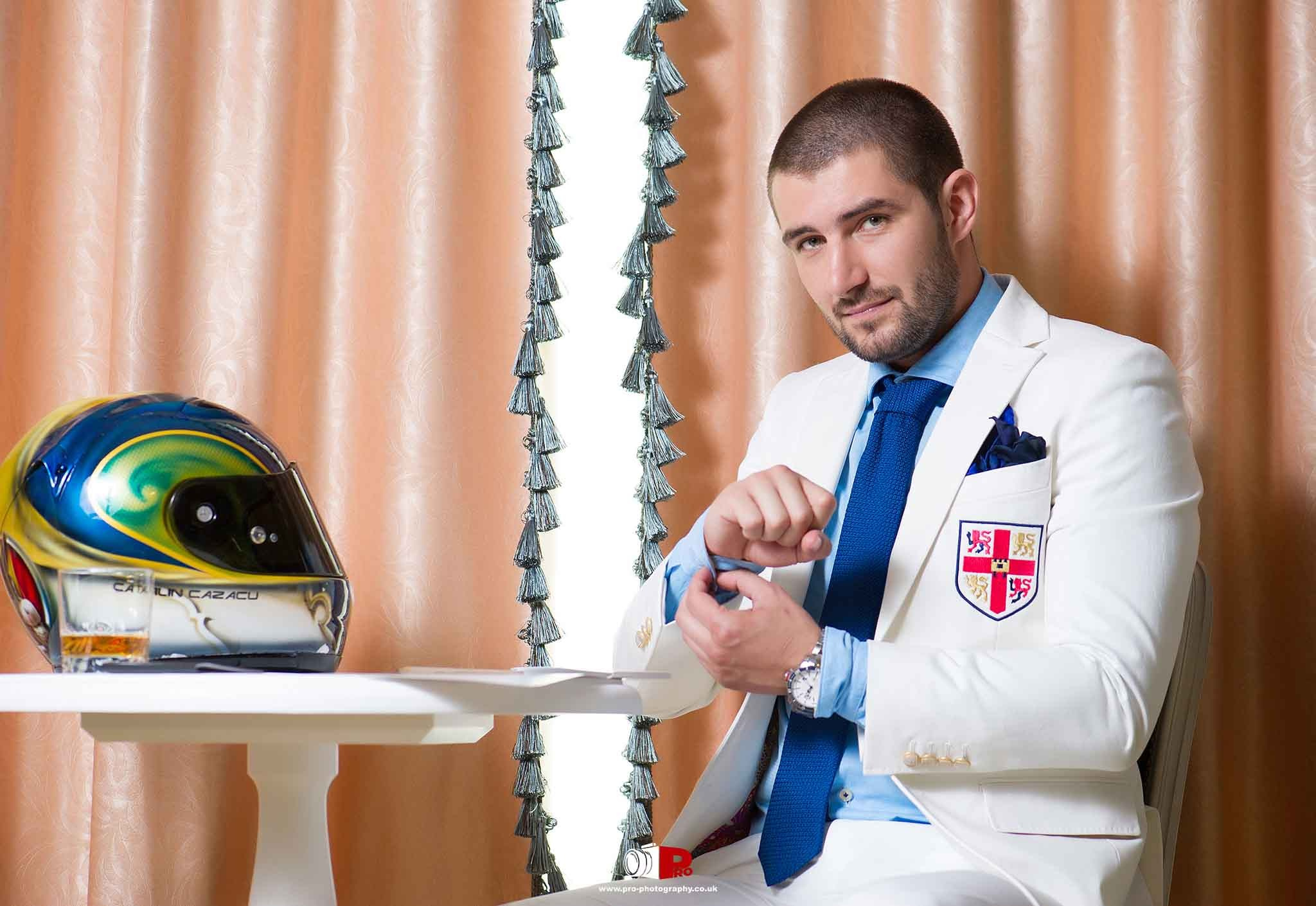 A man dressed in a white suit with a blue tie adjusts his cuffs while sitting beside a racing helmet on a table.
