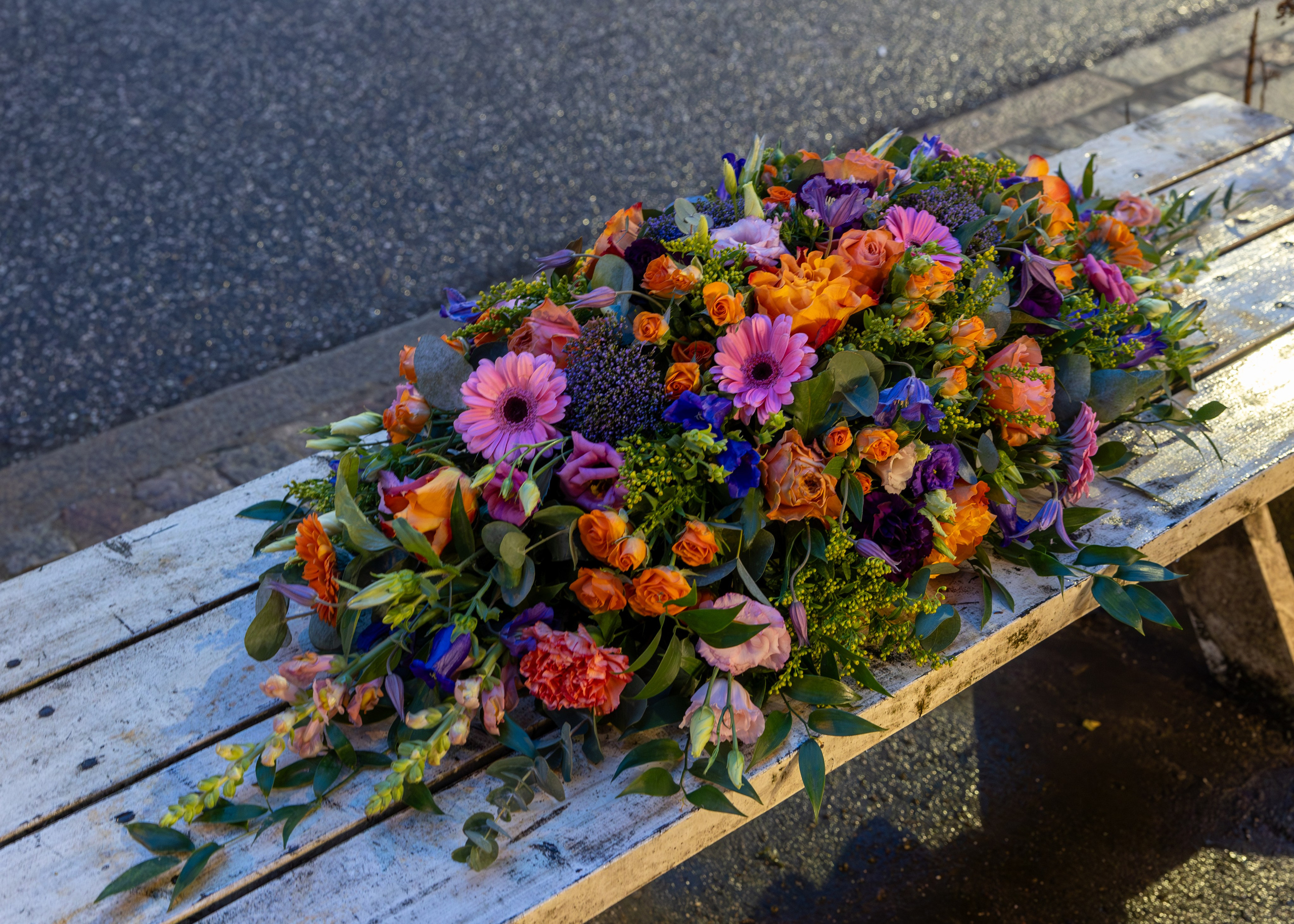 Kistepynt - Et eksklusivt farvel i hvid Farverig kistepynt med et let og levende udtryk. Blomsterstilkene har længde og højde, og fylder flot på kistelåget. Farverne er flot sammensat i stærke farver fra de orange toner over i pink-lilla nuancer. Kistepynten vil sætte farve på ethvert kirkerum. 