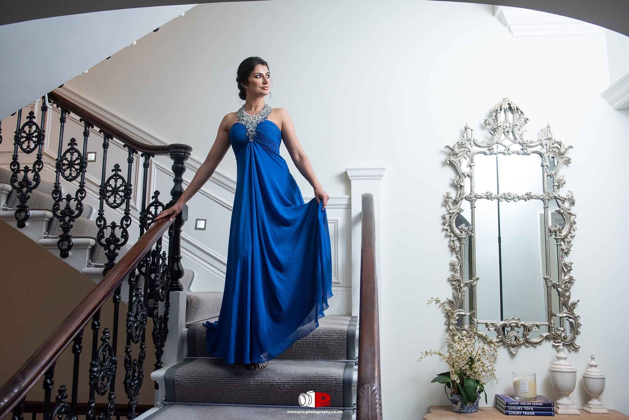 A sophisticated woman in a royal blue gown gracefully walks down a staircase in an elegant interior.