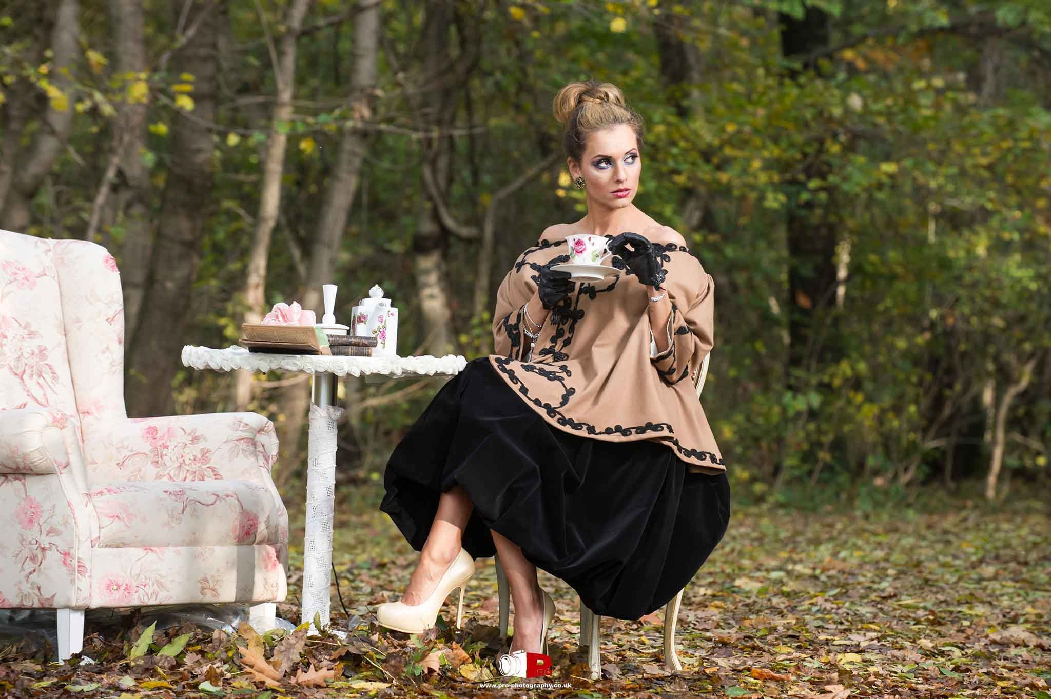 A stylish woman in a vintage-inspired outfit sipped tea in a serene forest beside a floral armchair and tea set.