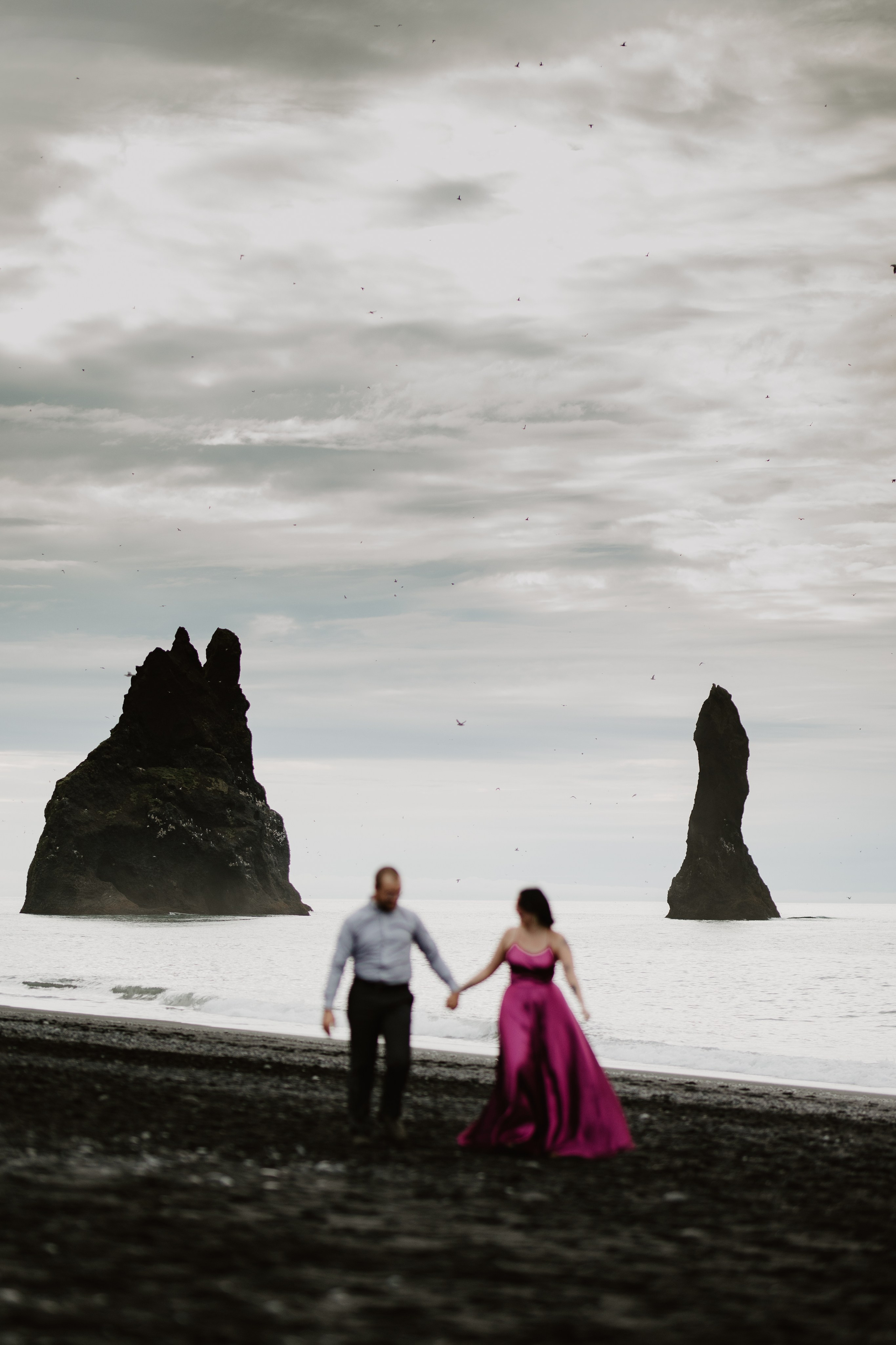 Love and adventure—couple walking along Reynisfjara Beach, Iceland, with the Reynisdrangar sea stacks in the distance.