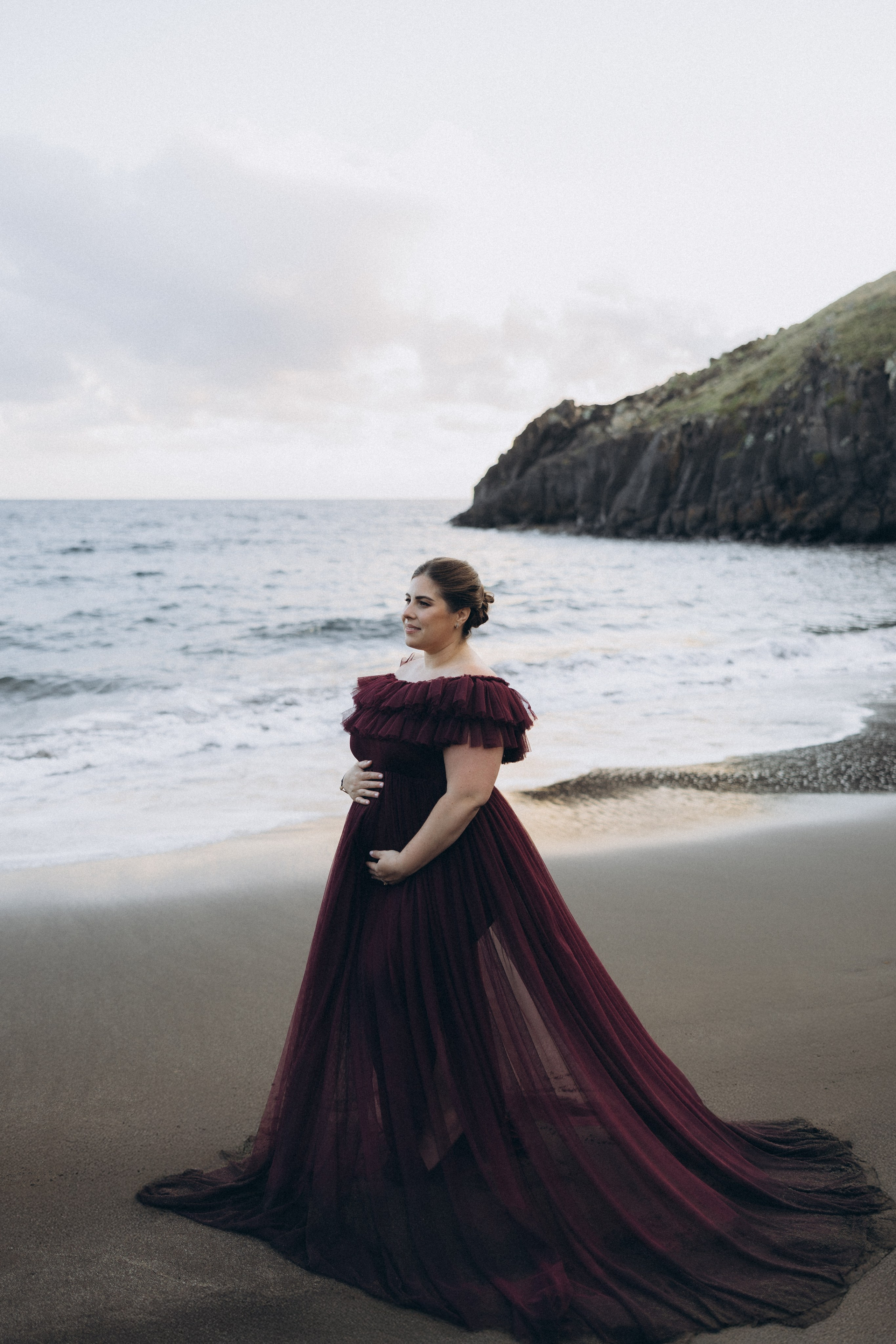 A glowing expectant mother standing on a cliff overlooking the ocean in Madeira, her dress flowing gently in the wind as the golden sunset casts a warm glow.