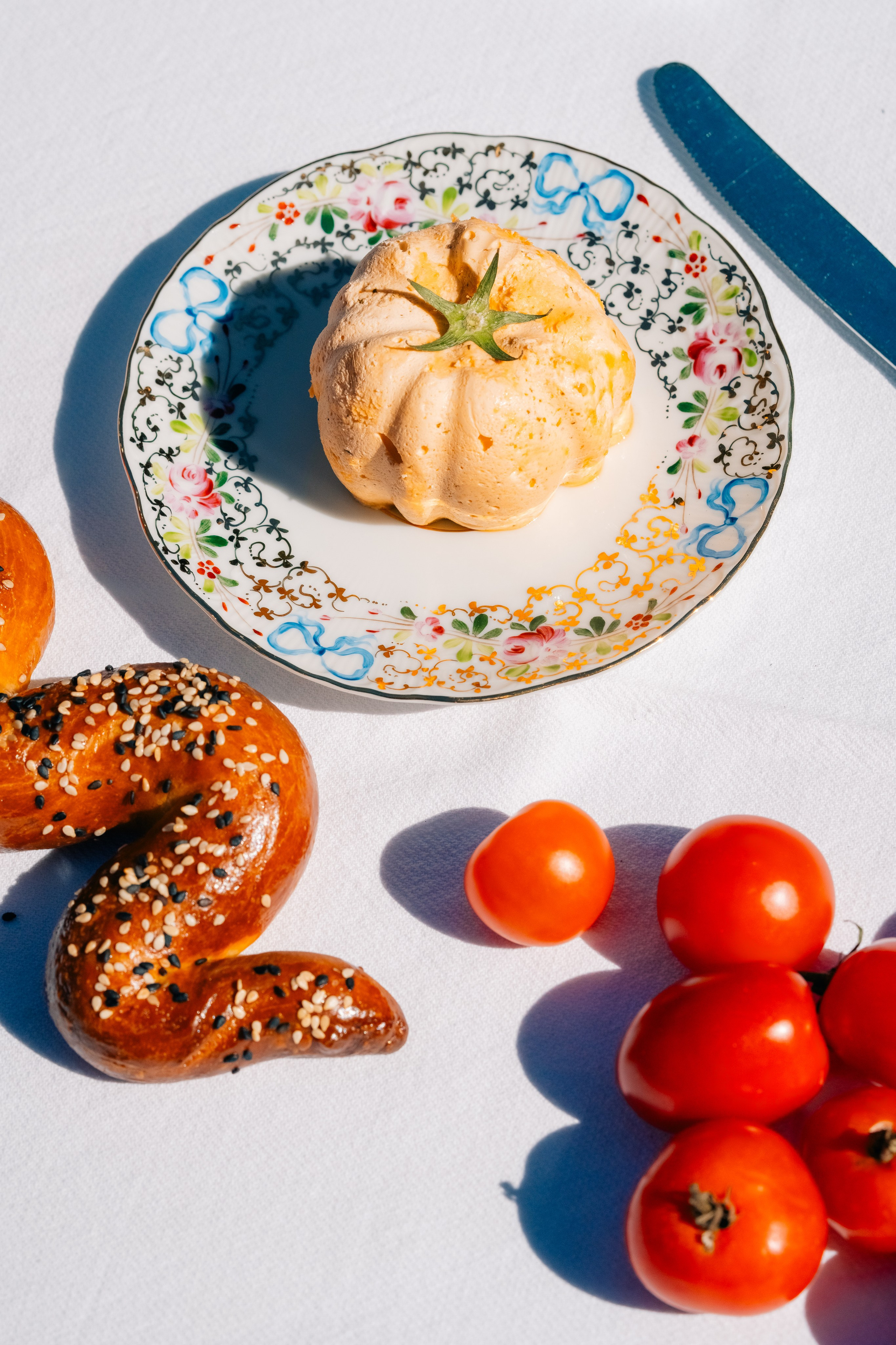 Arranjo de comida colorida com pastelaria, tomate e pão torcido. Uma mesa vibrante com um bolo num prato decorativo, tomates cereja frescos e um toque de pão com cobertura de sésamo.