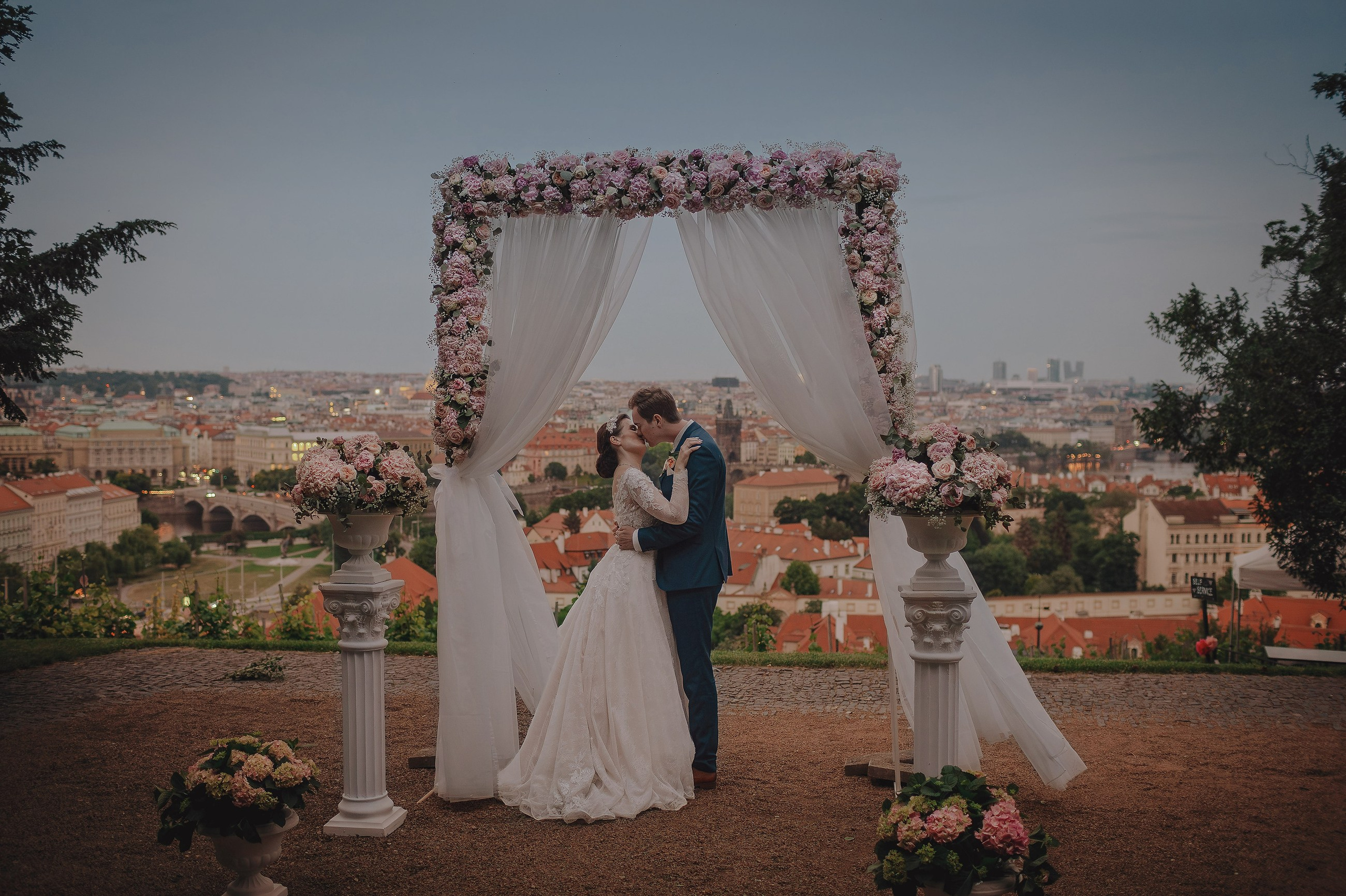 Newlyweds stand underneath a designer floral arch created in their honor as the twinkling lights of Prague is seen below them.