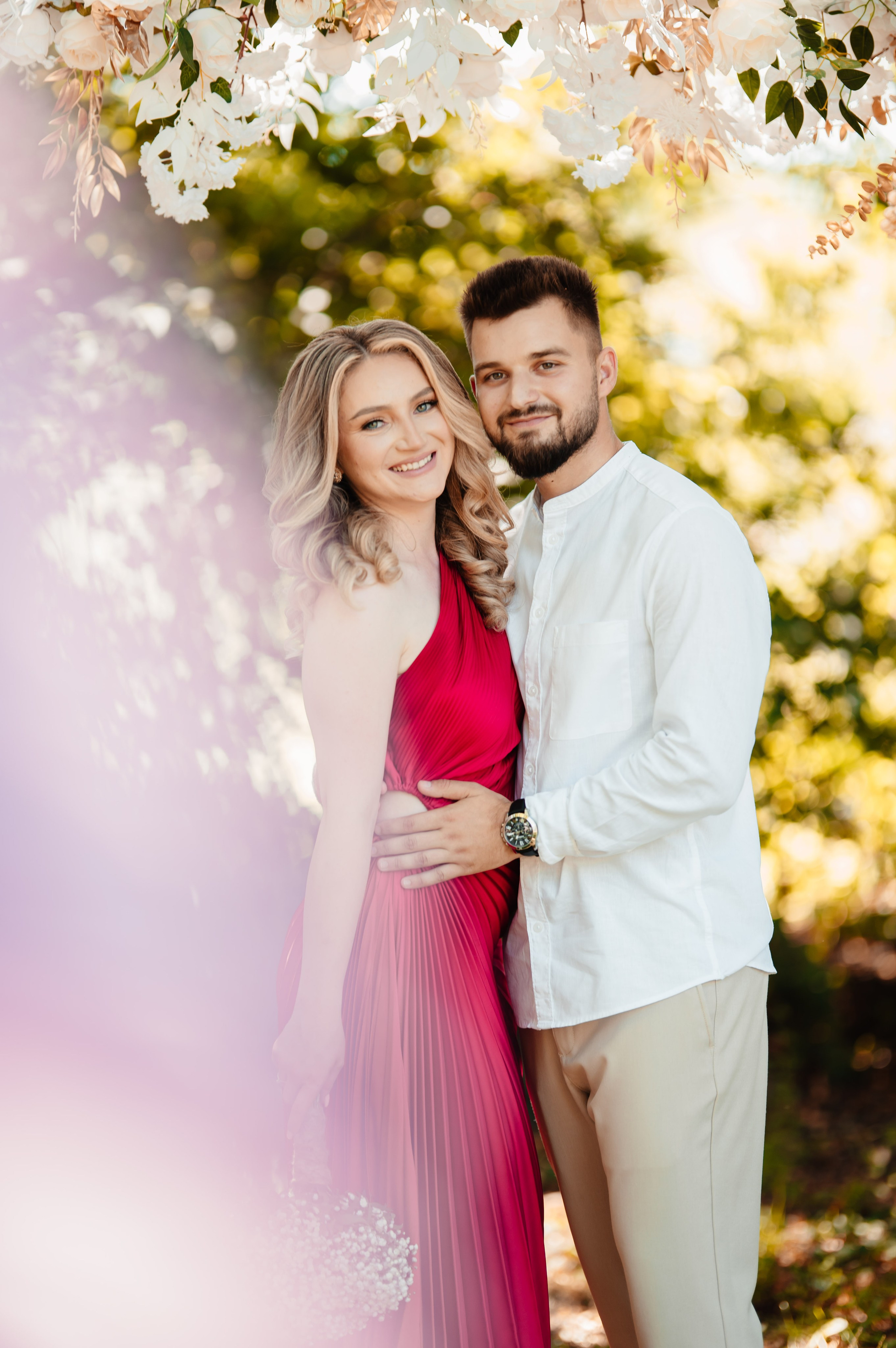 Cuplu zâmbitor îmbrățișat sub flori, într-un cadru romantic de primăvară.Dragoste înfloritoare: un cuplu radiant surprins într-un moment tandru, în natură.El în alb, ea într-o rochie elegantă roșie — un portret romantic sub ramuri înflorite.Priviri calde și îmbrățișări sub cerul senin — iubirea surprinsă în cea mai frumoasă lumină.Cuplu fericit într-un decor floral, radiind dragoste și armonie.