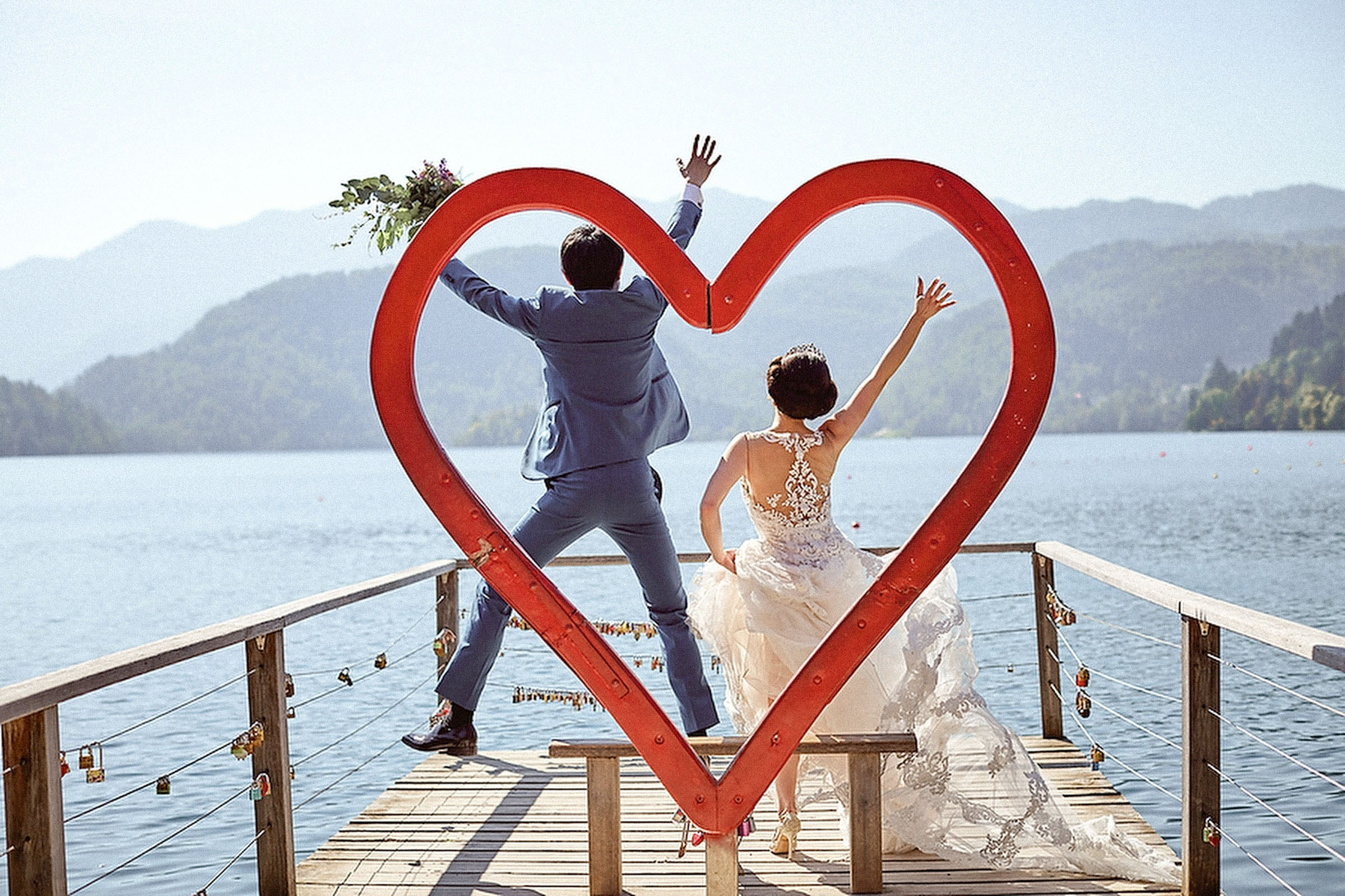 A groom wearing a blue suit and holding a bouquet and his bride is seen from behind as they jump in the air as seen through the 'Heart of Bled' 