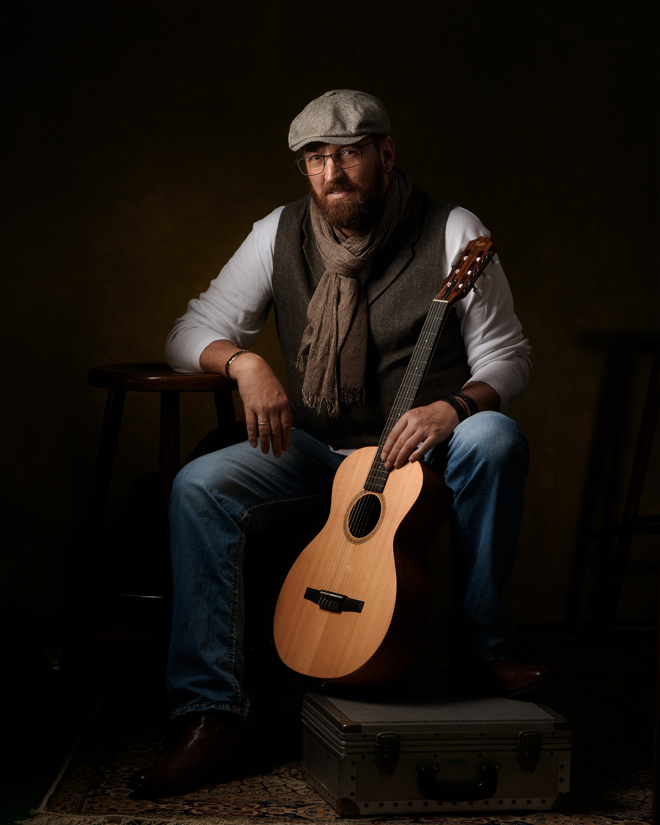 Er ist ein Musiker mit Leidenschaft und Herz. Er sitzt entspannt auf einem Stuhl und hält seine Gitarre mit der linken Hand