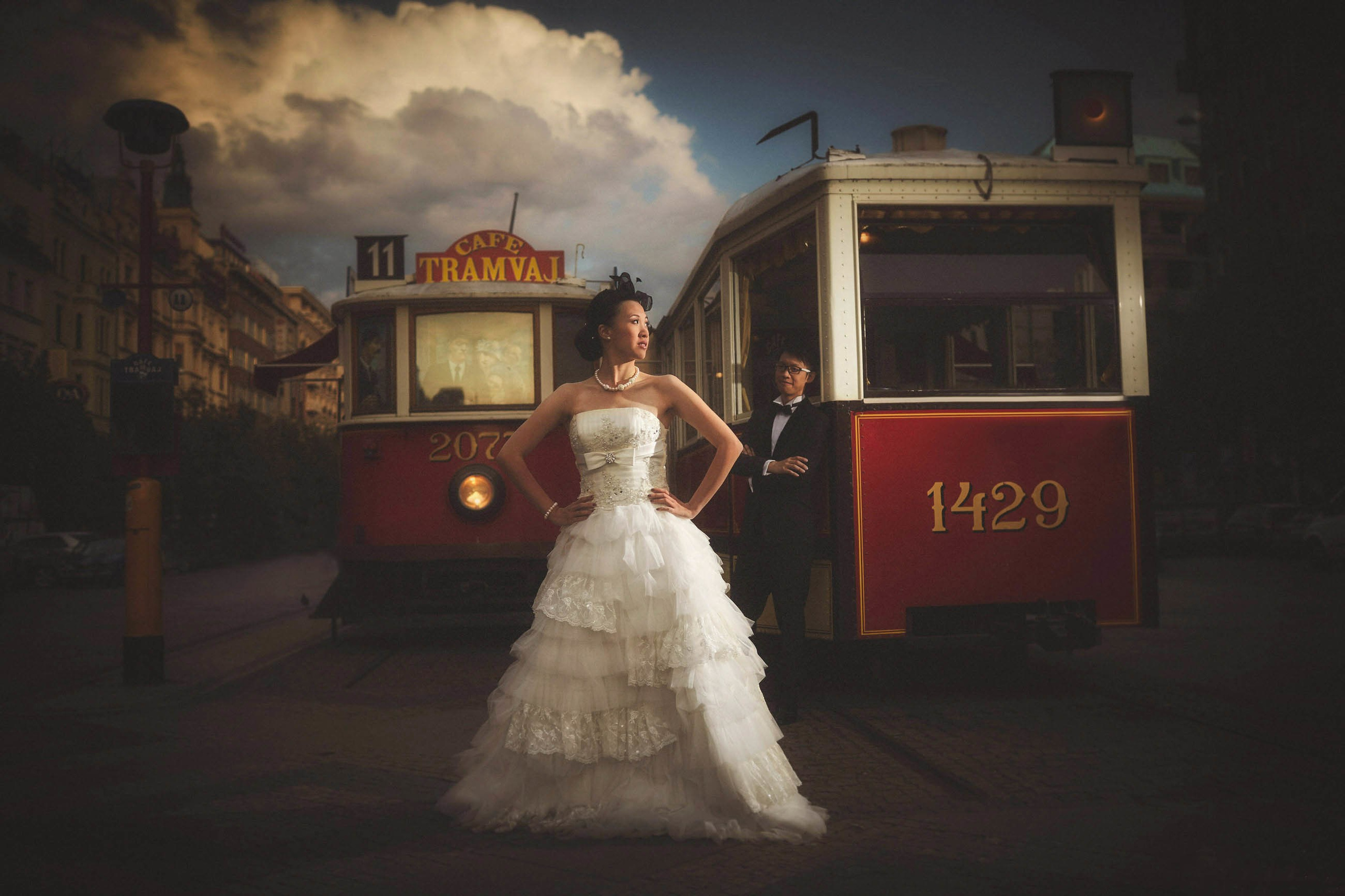 Chic Hong Kong bride with a fashionable headpiece and her groom posing near the vintage trams of Prague, embodying a cultural fusion.