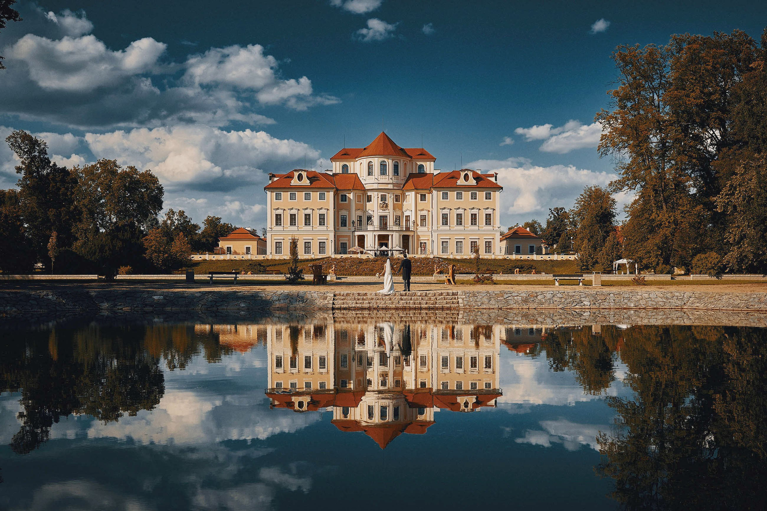 As the deep blue sky is broken by puffy, white clouds, a bride and groom are reflecting in the waters of a man made lake as they view the opulent Chateau Liblice, a 17-th century masterpiece in Czechia.