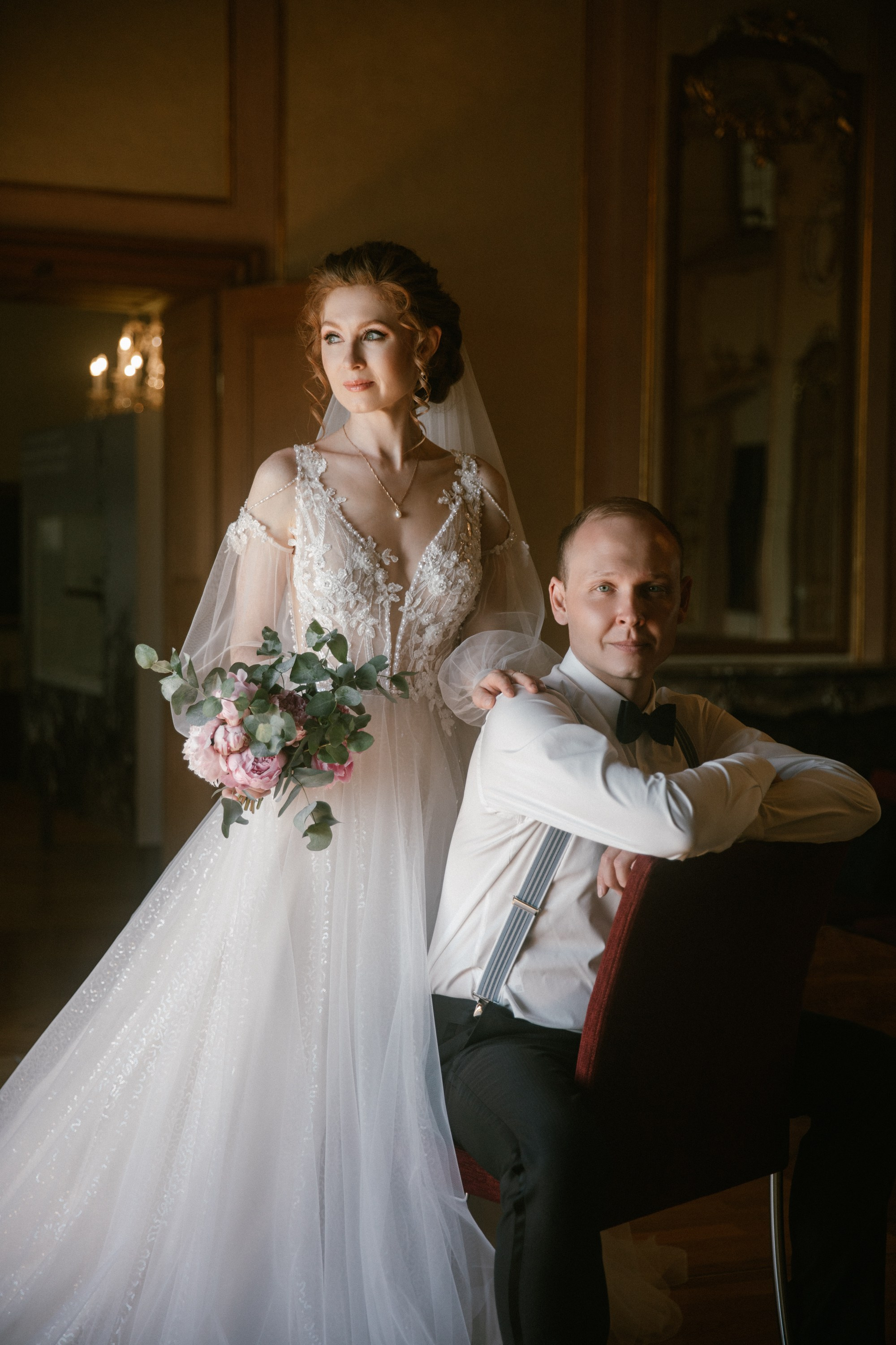 Bride with bouquet stands beside seated groom in the Mirror Hall of Schloss Meersburg