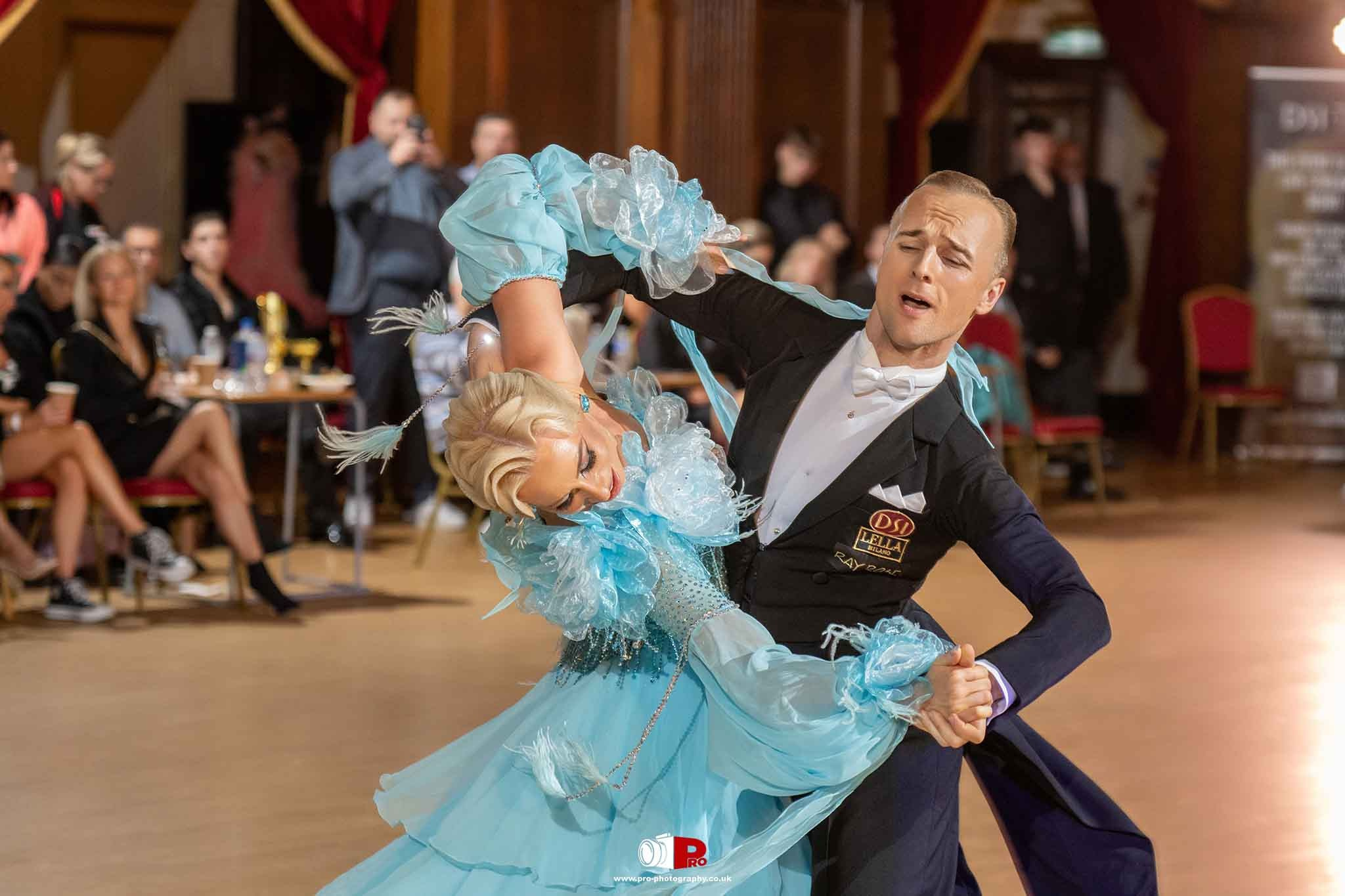 A ballroom dance couple elegantly performing in a graceful blue gown with feathered sleeves in a dance competition.