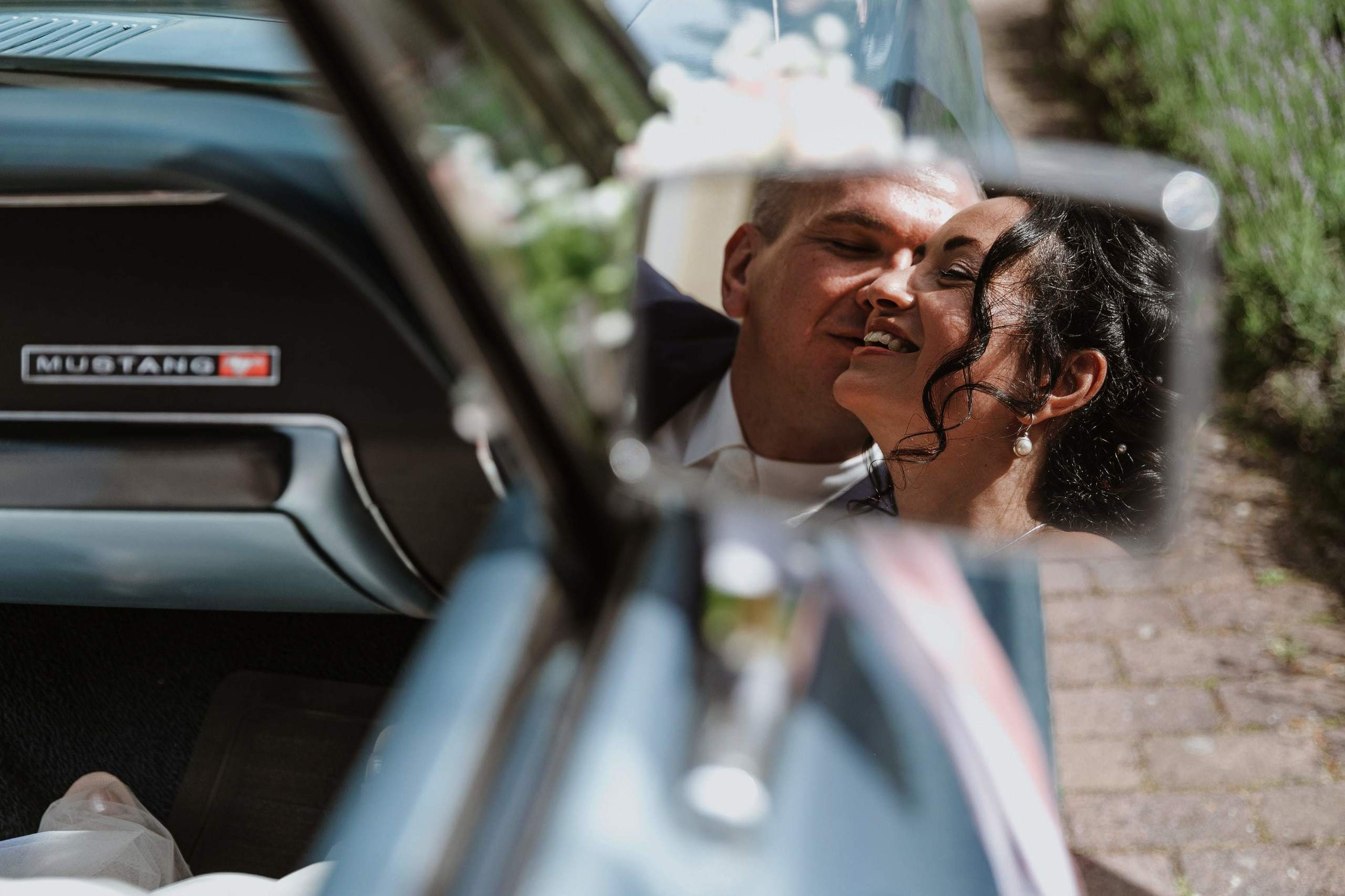 Der Bräutigam küsst die Braut auf die Wange, ihr Spiegelbild ist im Rückspiegel des Mustang-Cabriolets sichtbar.
