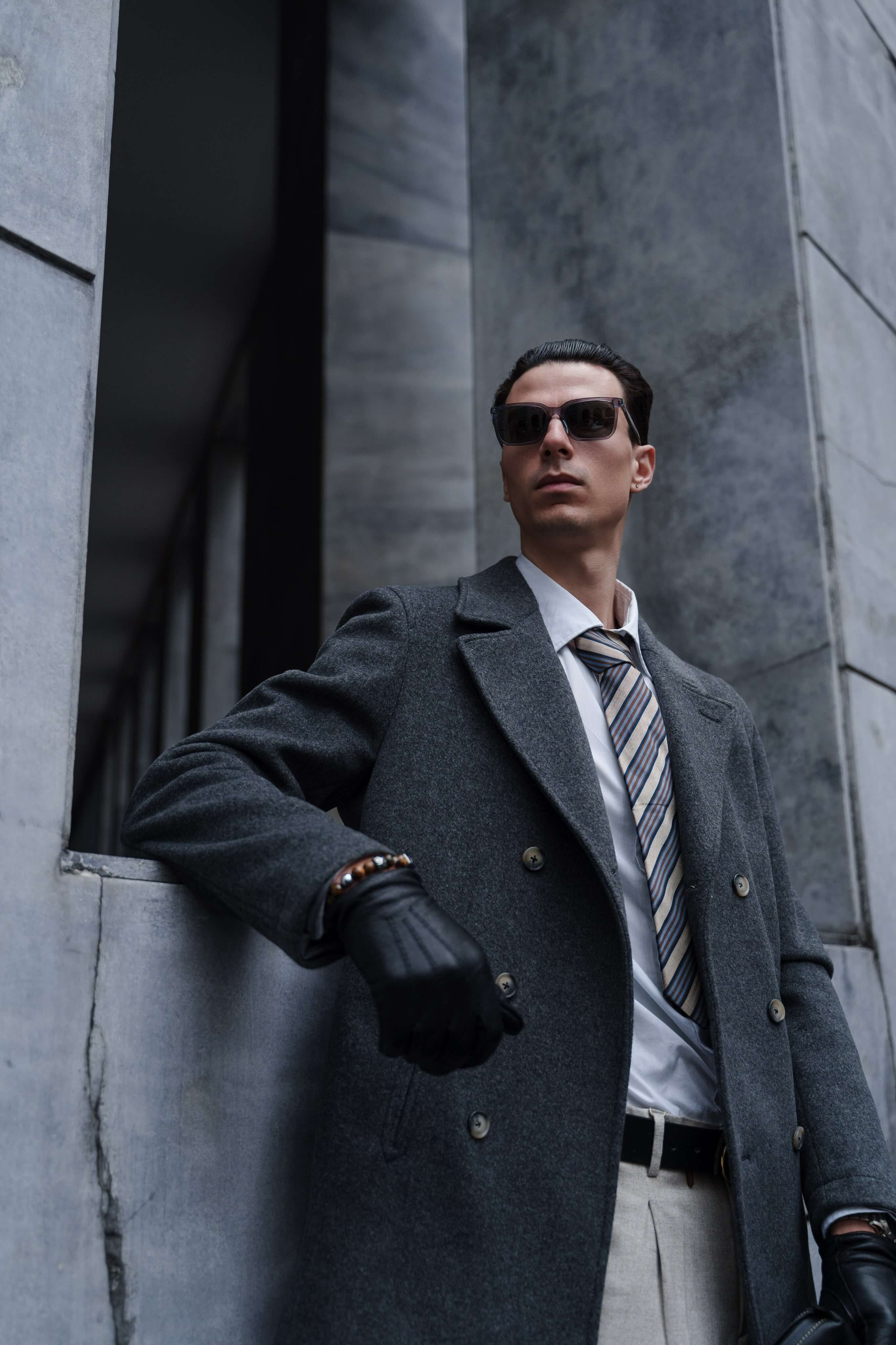 Man in dark sunglasses and grey wool coat leaning against modern marble wall