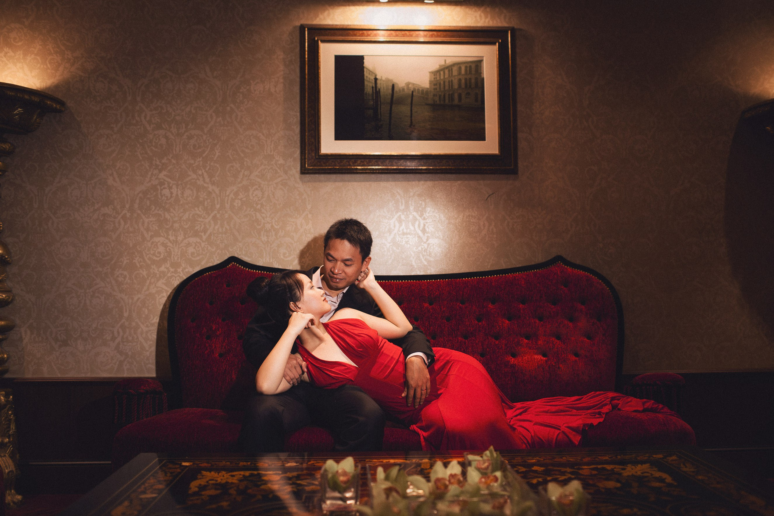 A sexy Woman in a tight red dress reclines on her fiancee's lap on antique chair at the historic Hotel Danieli in Venice.