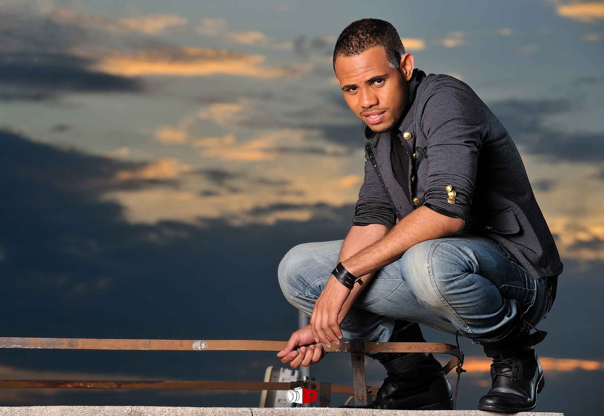 A fashionable man crouching on a rooftop with a sunset sky in the background.