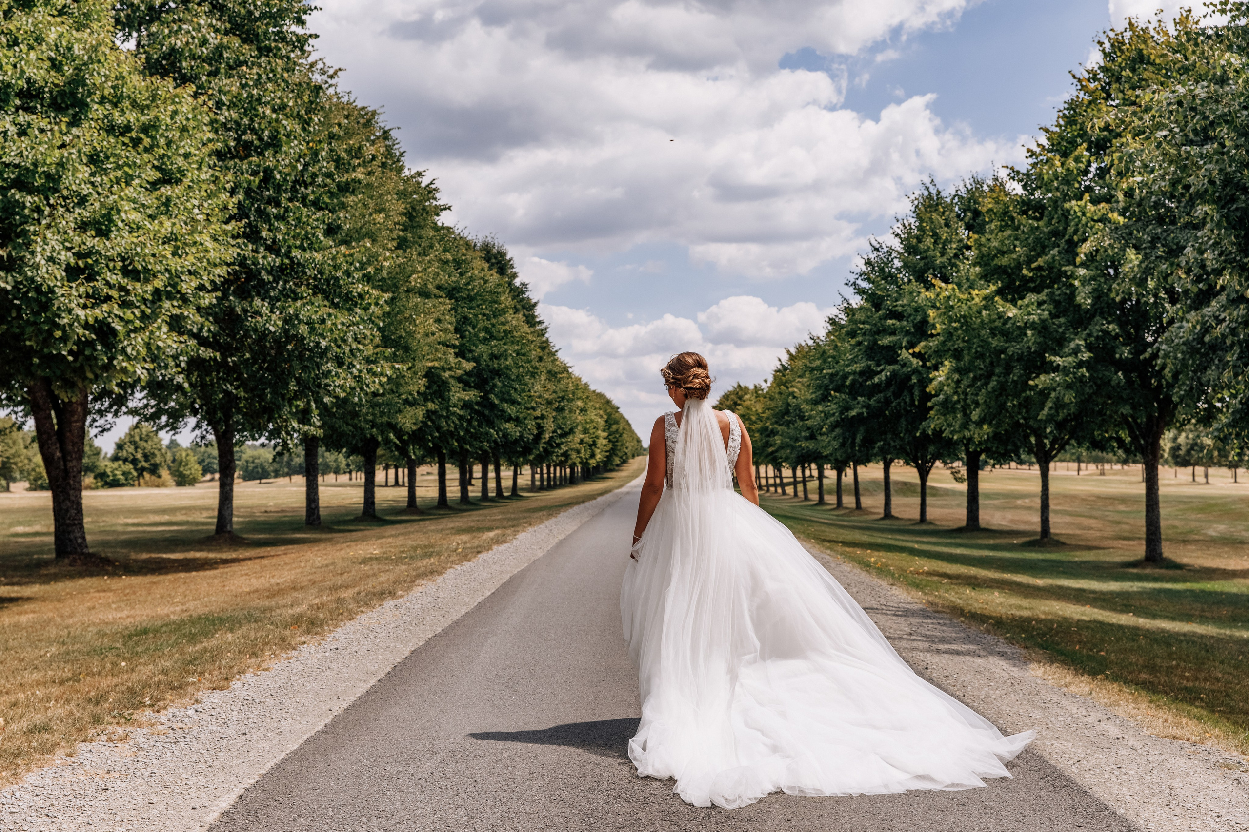 Die Braut geht durch eine von Bäumen gesäumte Allee, ihr Kleid weht elegant im Wind.