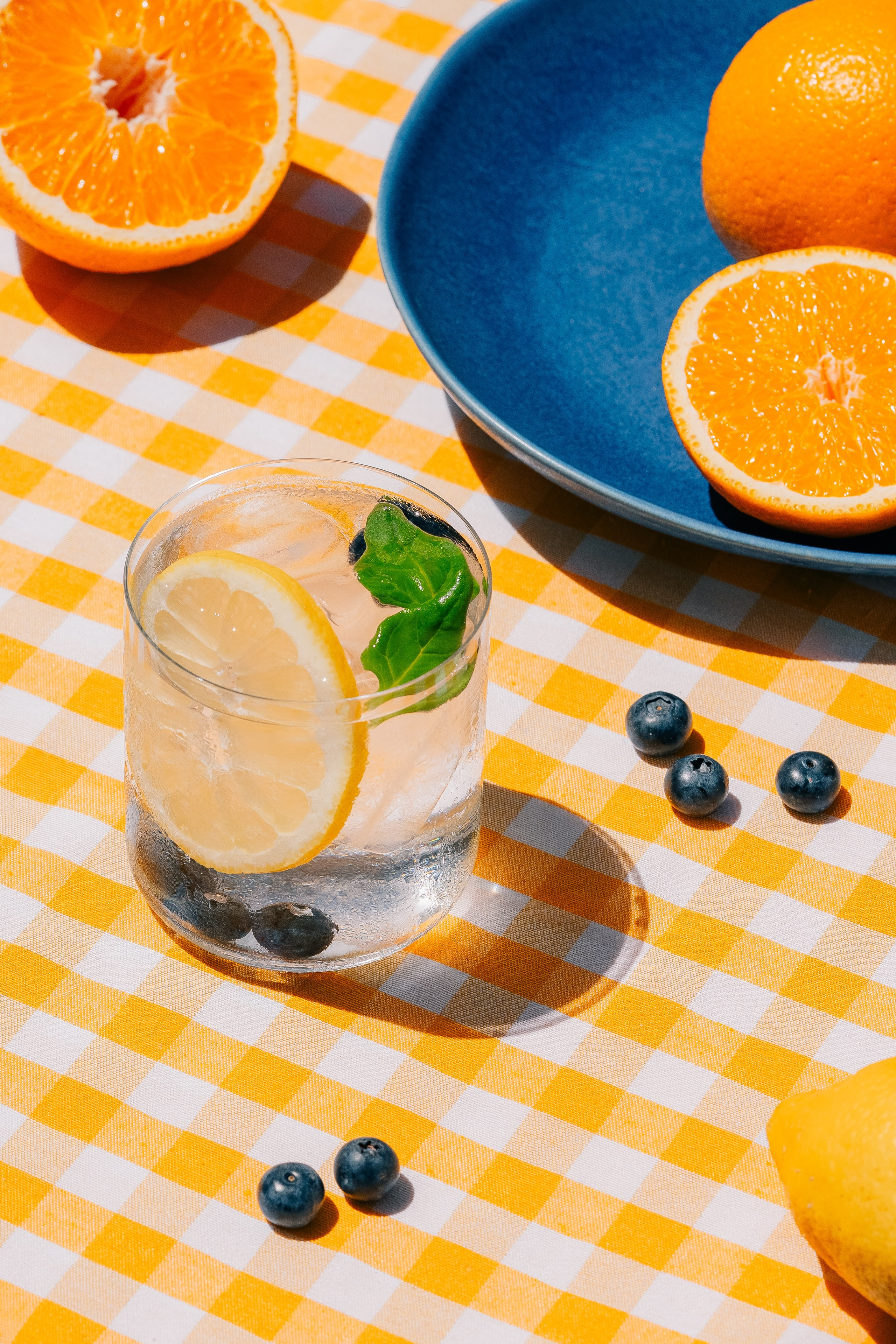 Refrescante bebida de limão e mirtilo em toalha de mesa quadriculada. Um refrescante copo de bebida de limão e mirtilo em uma toalha amarela xadrez, guarnecida com ervas frescas. Colocado perto de prato azul servido com laranjas.