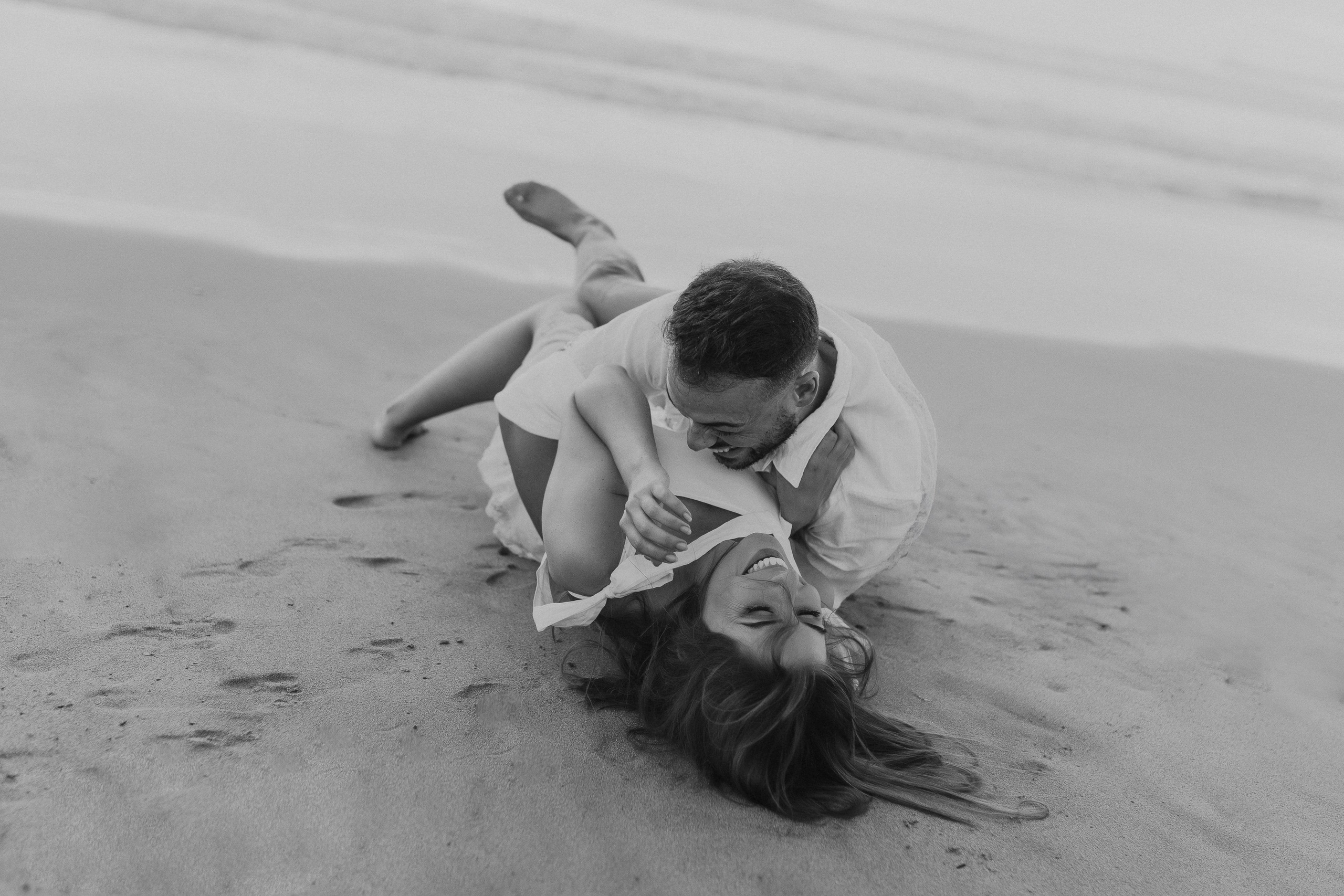 Casal ri e brinca deitado na areia durante sessão de noivado espontânea na praia do norte de Portugal.