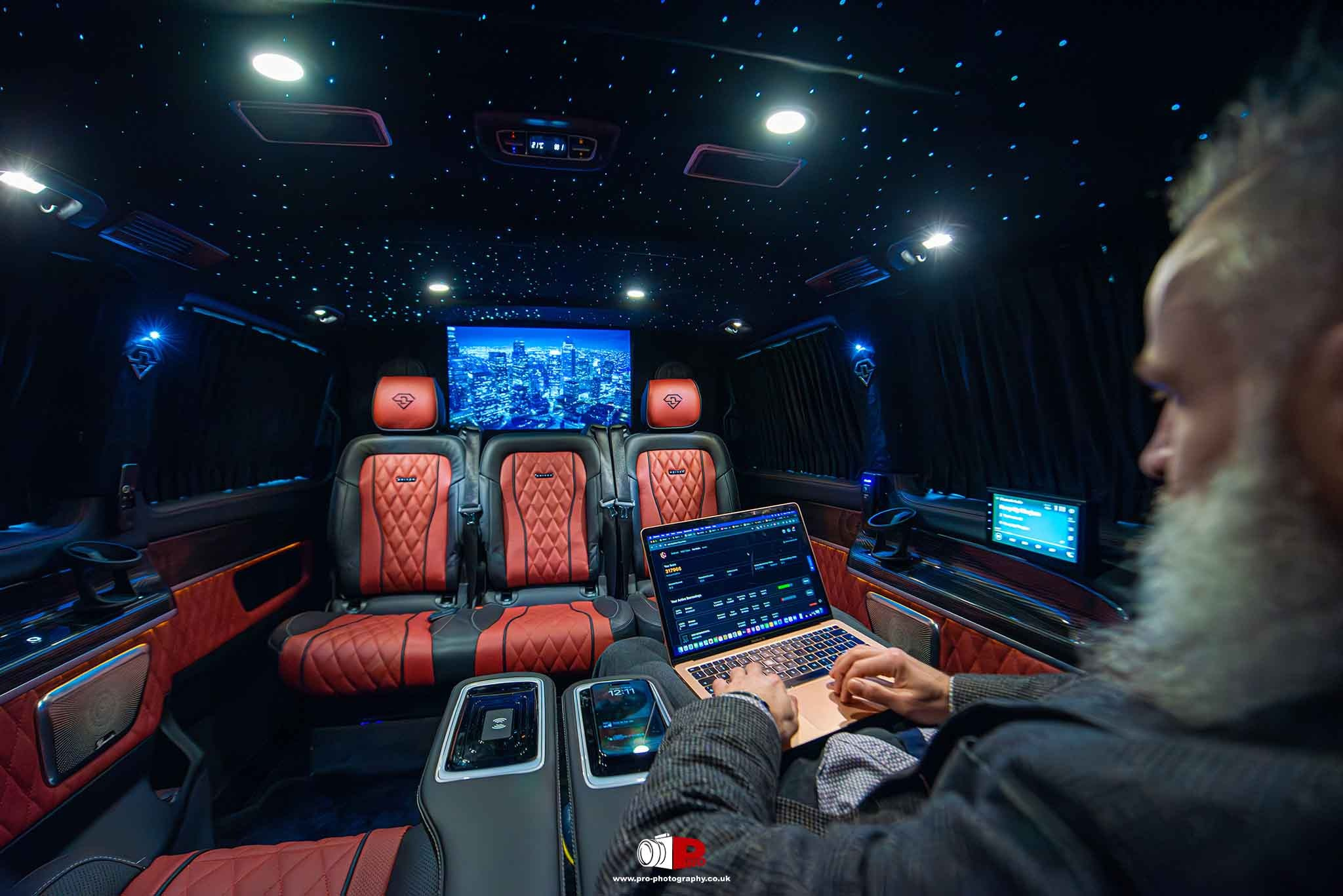 A luxurious van interior featuring plush red seats, a ceiling starry light effect, and a man working on a laptop.