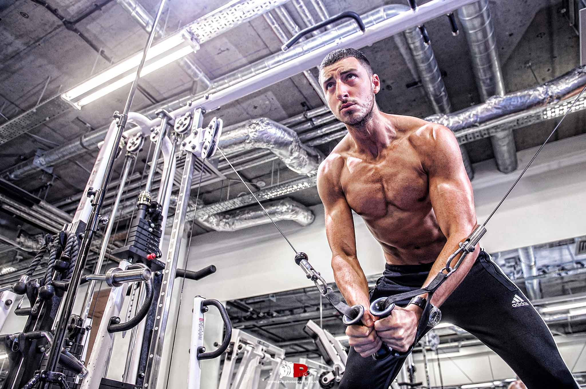 A muscular male model intensely performing a cable crossover exercise in a modern industrial-style gym.