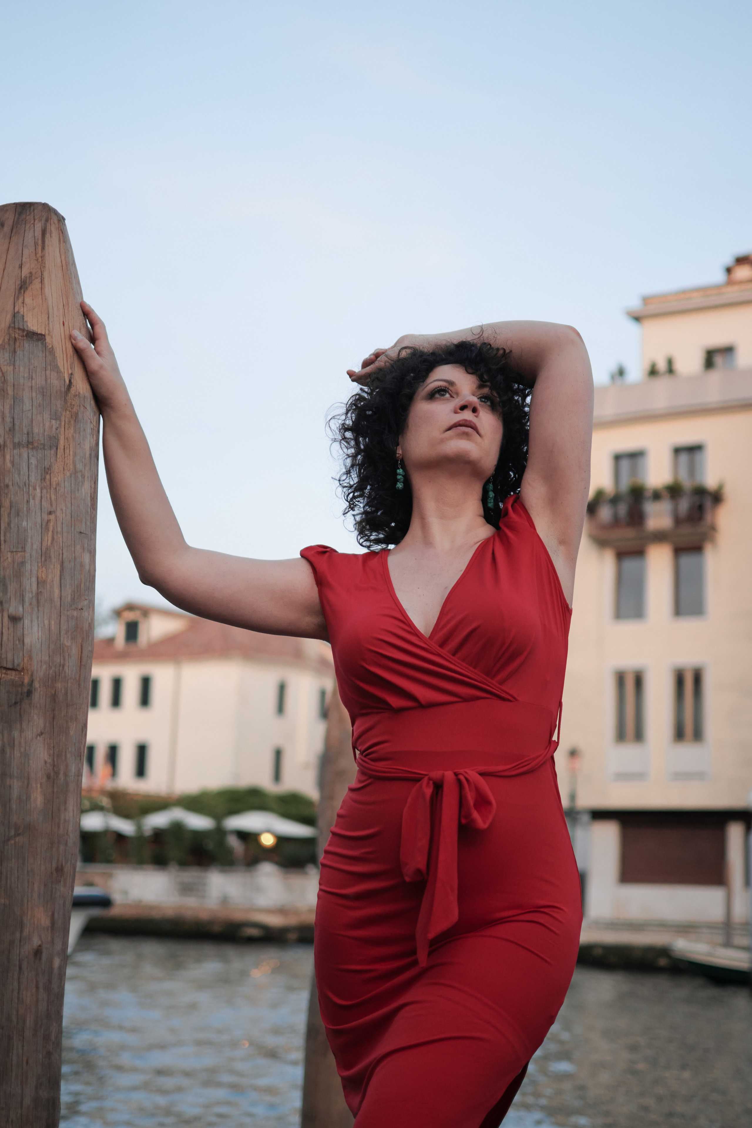 A photographer in Venice, Italy taking portrait photos of a womanwith red dress. The woman is standing in front of a beautiful bridge with the iconic canals of Venice in the background. The photographer is crouching down with a camera in hand, capturing girl's pose and expression.