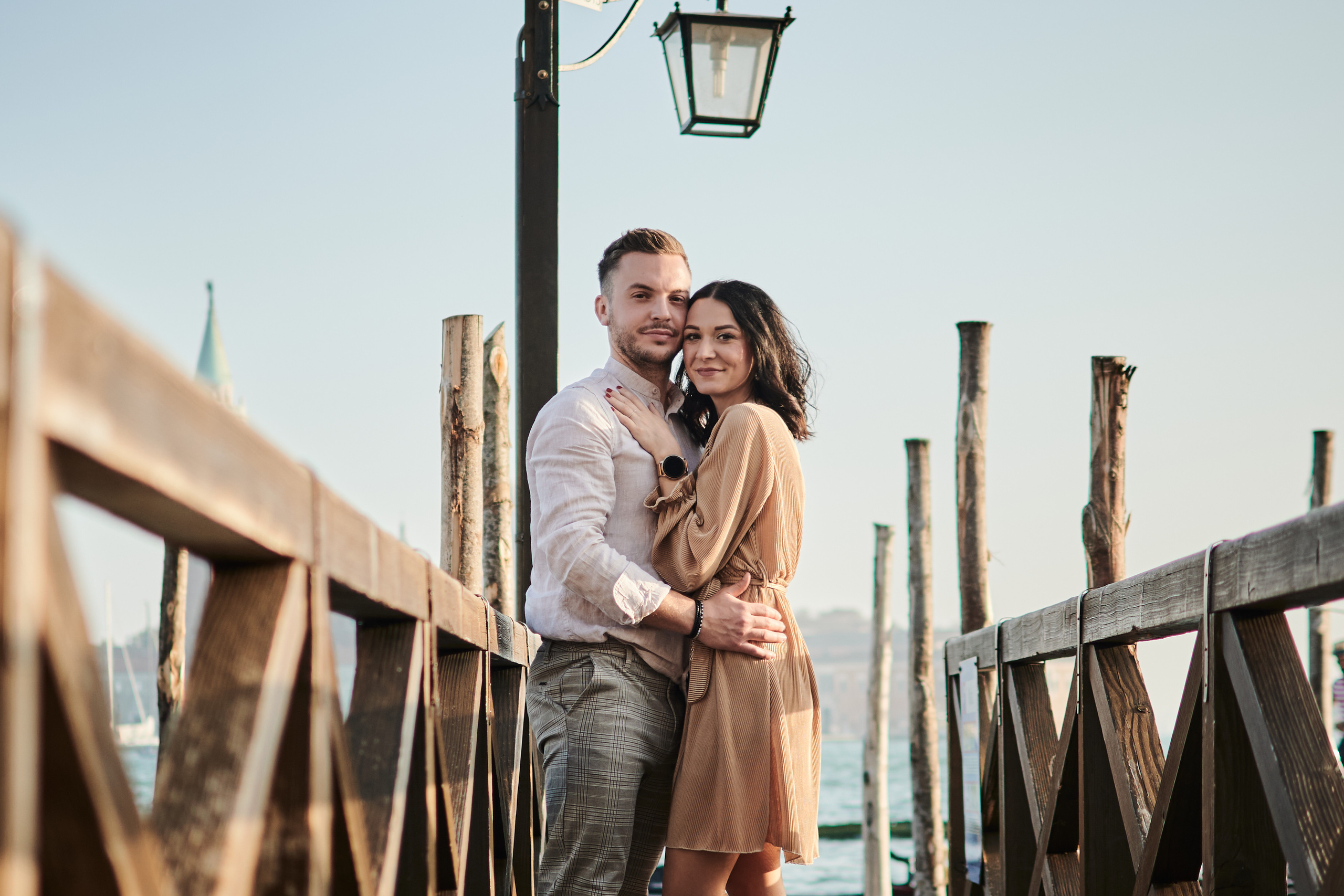 A couple stands hand in hand, gazing out at the breathtaking view of the Laguna and San Marco square in Venice. The warm hues of the sunset reflect off the water, casting a romantic glow on the scene. The couple's love is evident in their tender embrace, as they share a moment of pure bliss amidst the stunning panorama. The iconic columns of San Marco and San Todaro tower behind them, offering a timeless backdrop to their love story