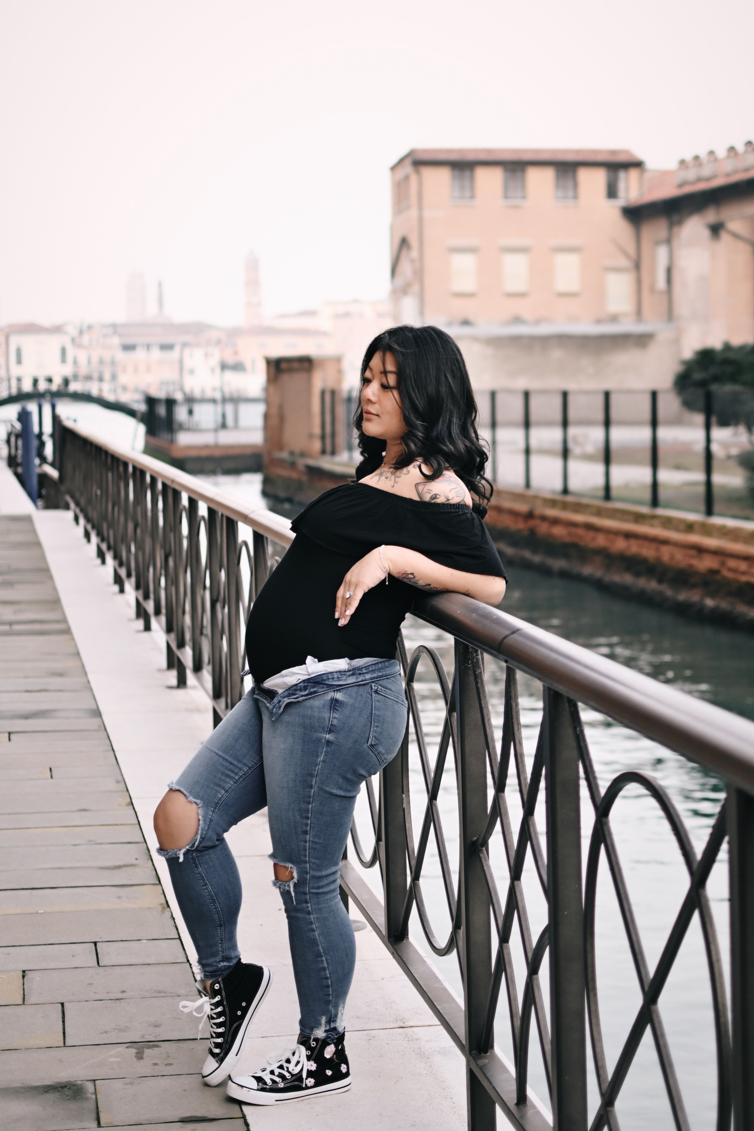 A pregnant young woman stands on a bridge in Venice, wearing a pair of comfortable jeans and a black blouse. she gazes out at the bustling canal below. The sunlight glimmers on the water, casting a warm glow over the scene. In the background, ancient buildings and colorful boats lend a sense of history and romance to the setting. The woman's pose is relaxed yet confident, conveying her excitement and anticipation for the new life growing inside her