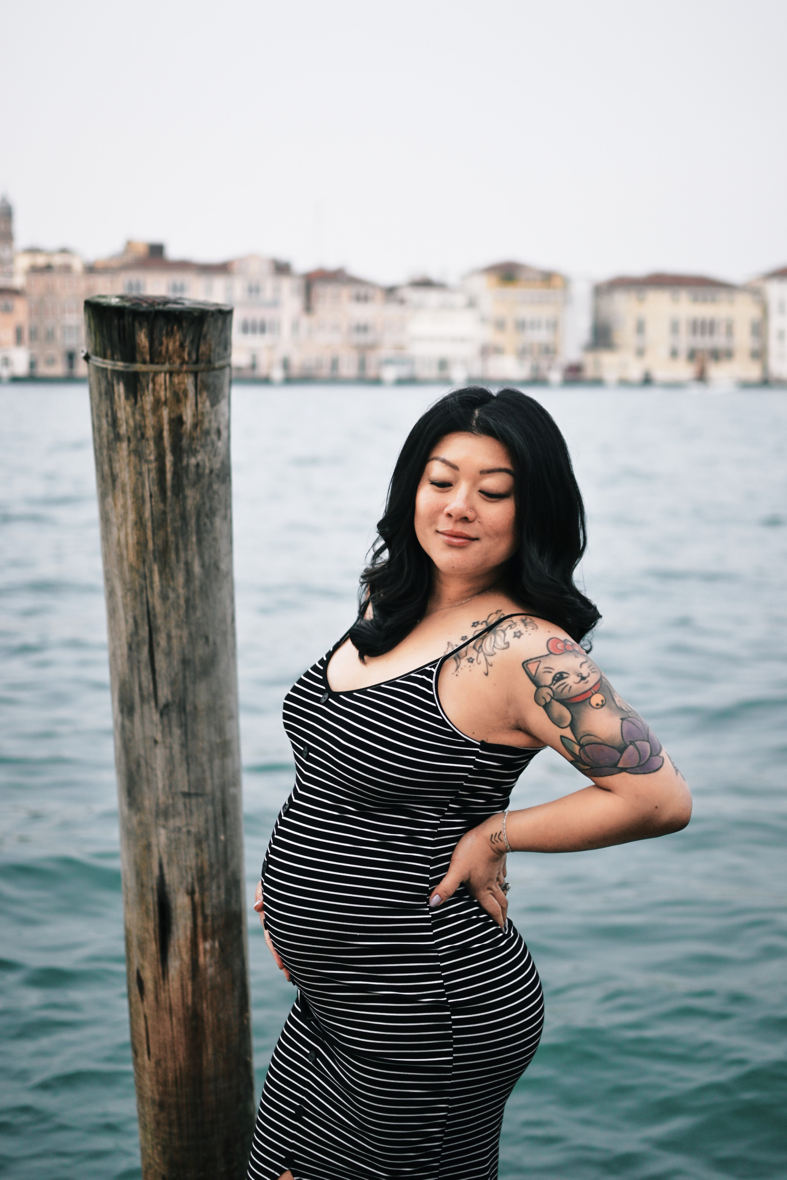 A glowing pregnant woman stands by the water's edge in Venice, Italy. She cradles her growing belly with one hand and gazes out at the stunning view of the city's canals and historic buildings. The soft light of the sun sets behind her, casting a warm glow on her face and highlighting the intricate lace of her dress. The quiet beauty of the moment is captured in this serene portrait, as the woman awaits the arrival of her precious little on