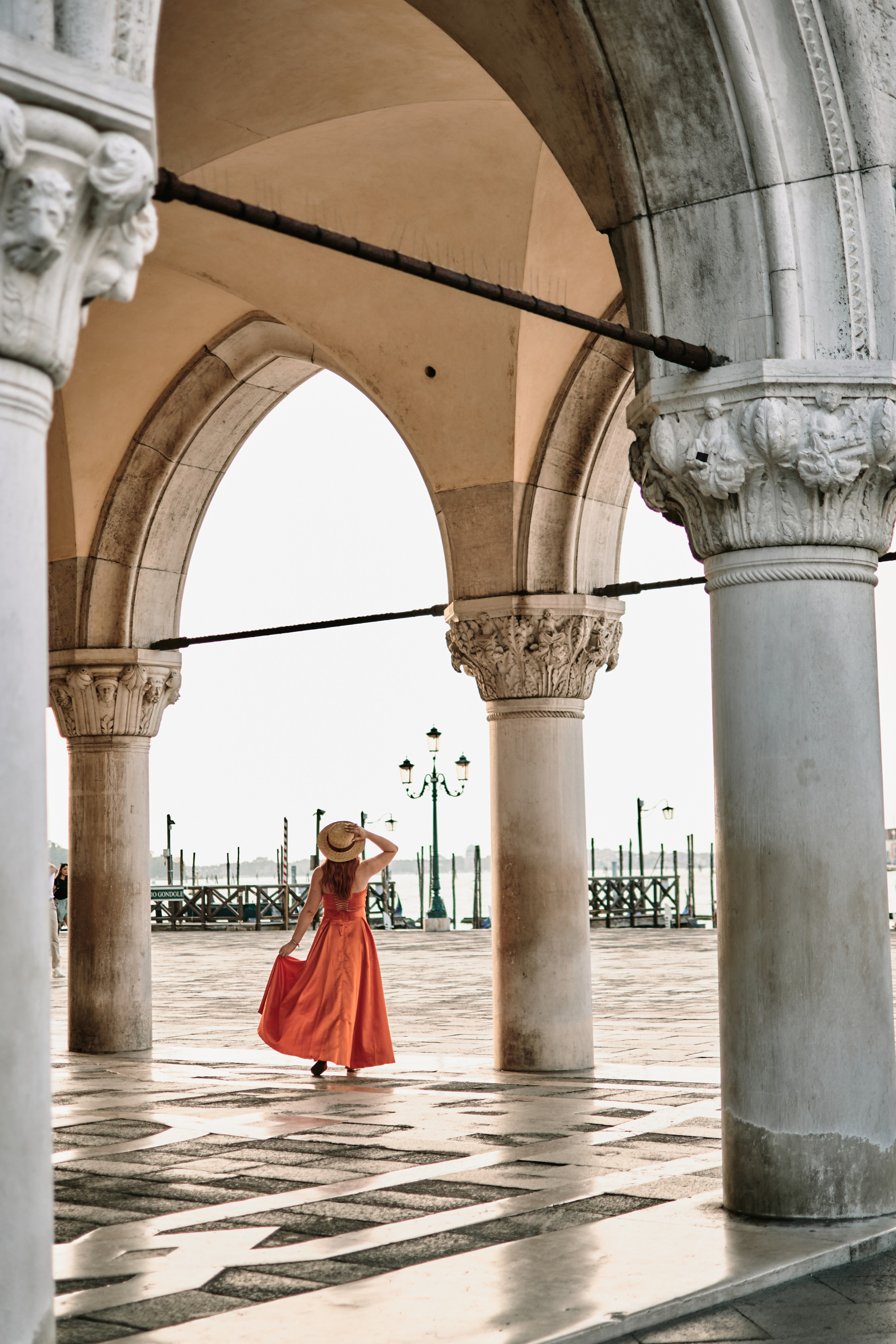 A stunning young woman in orange yellow dress stands in San Marco Square, Venice, with the historic Arca di San Marco in the background. The architectural details of the Arca and the surrounding buildings add to the romantic atmosphere of the scene. The woman's dress drapes elegantly around her figure, adding to the captivating allure of the photo. The contrast between the dark dress and the warm colors of the surrounding buildings adds to the overall visual impact of the image, creating a sense of mystery and intrigue.