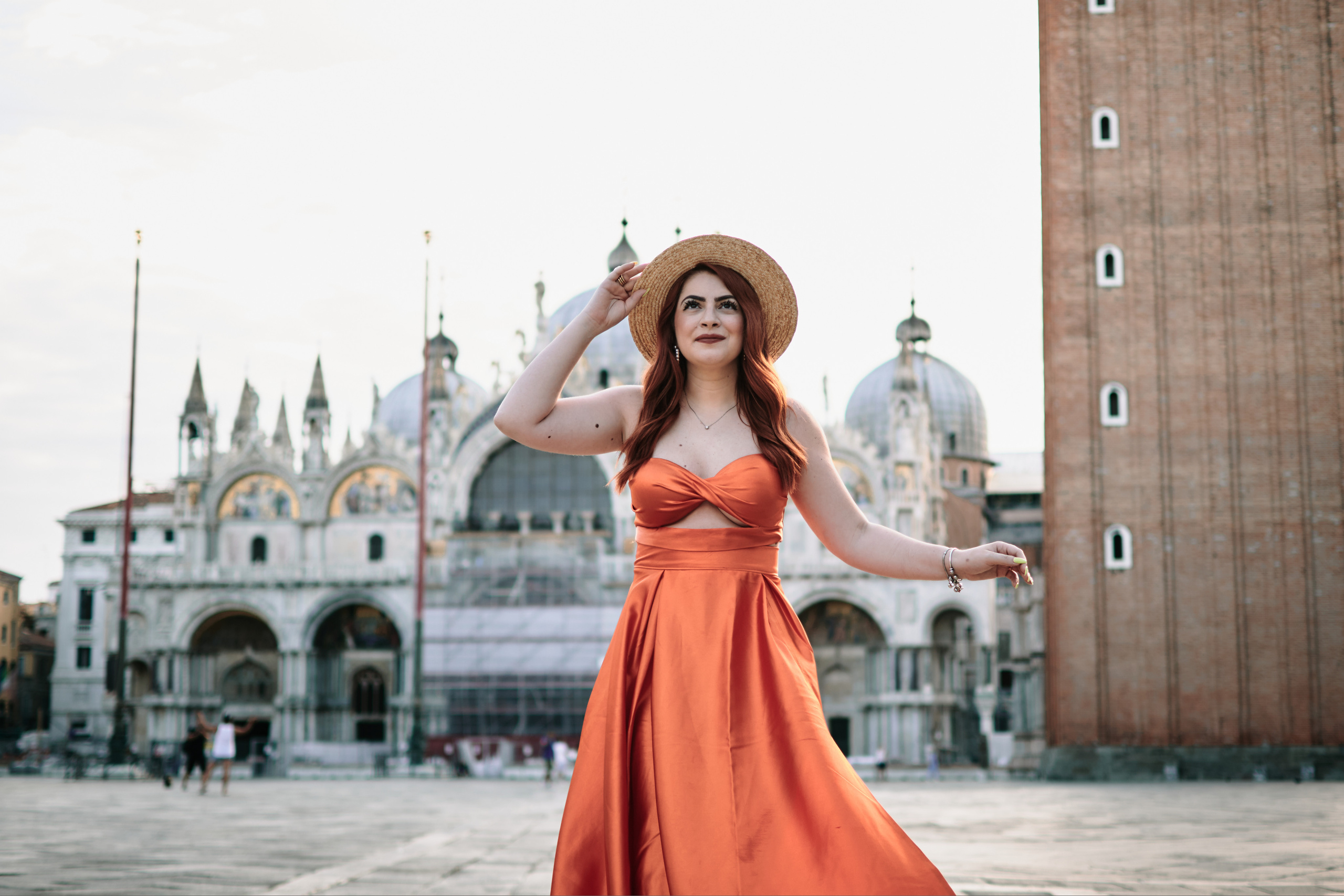 A young woman  stands on San Marco square in Venice. She wears a flowing orange sundress that complements the warm sunlight reflecting off the water behind her. The red and white striped poles of a gondola rest in the background, adding to the Italian charm of the scene. She looks relaxed and carefree, enjoying the beautiful day in this timeless city