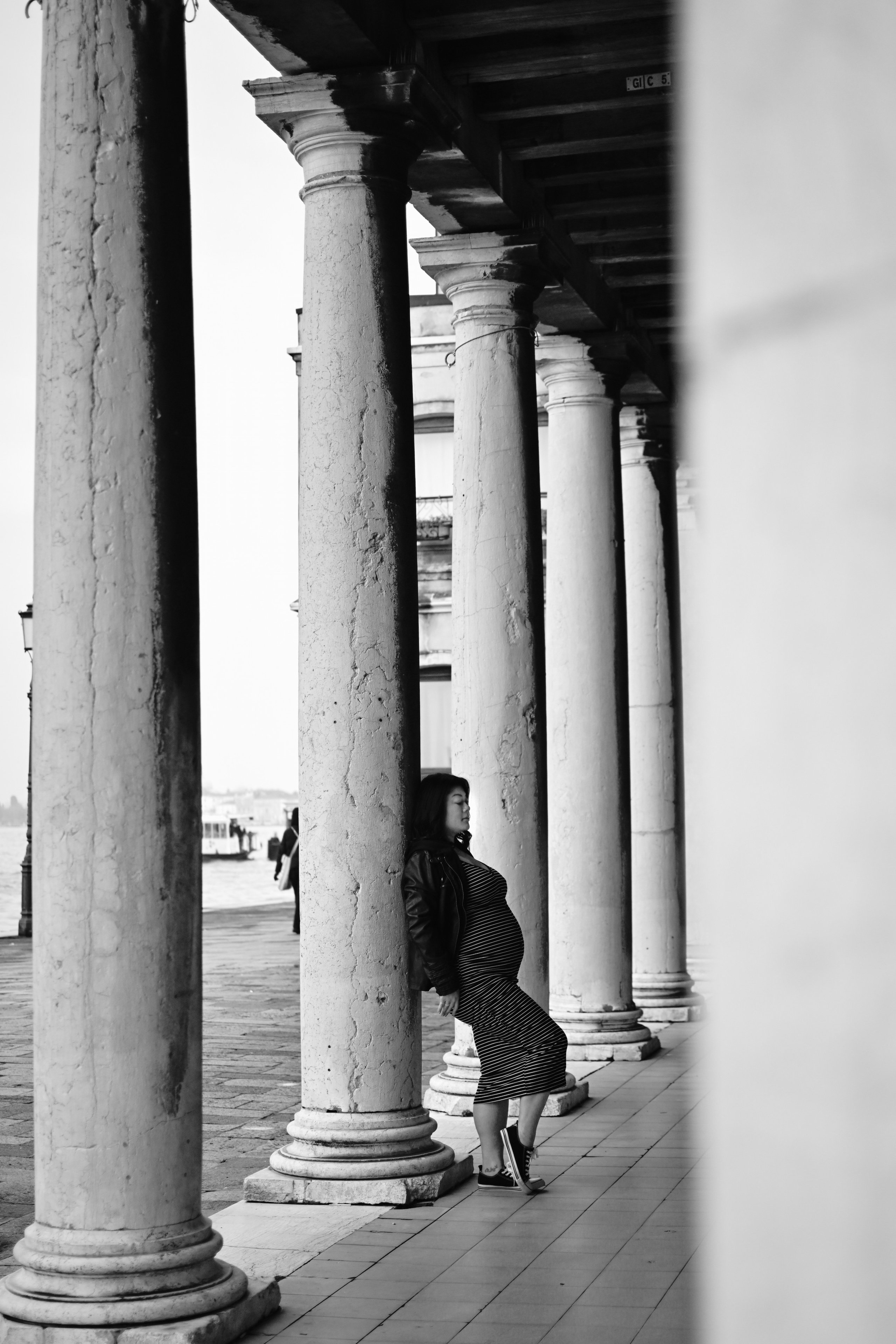 A glowing pregnant woman stands by the water's edge in Venice, Italy. She cradles her growing belly with one hand and gazes out at the stunning view of the city's canals and historic buildings. The soft light of the sun sets behind her, casting a warm glow on her face and highlighting the intricate lace of her dress. The quiet beauty of the moment is captured in this serene portrait, as the woman awaits the arrival of her precious little one