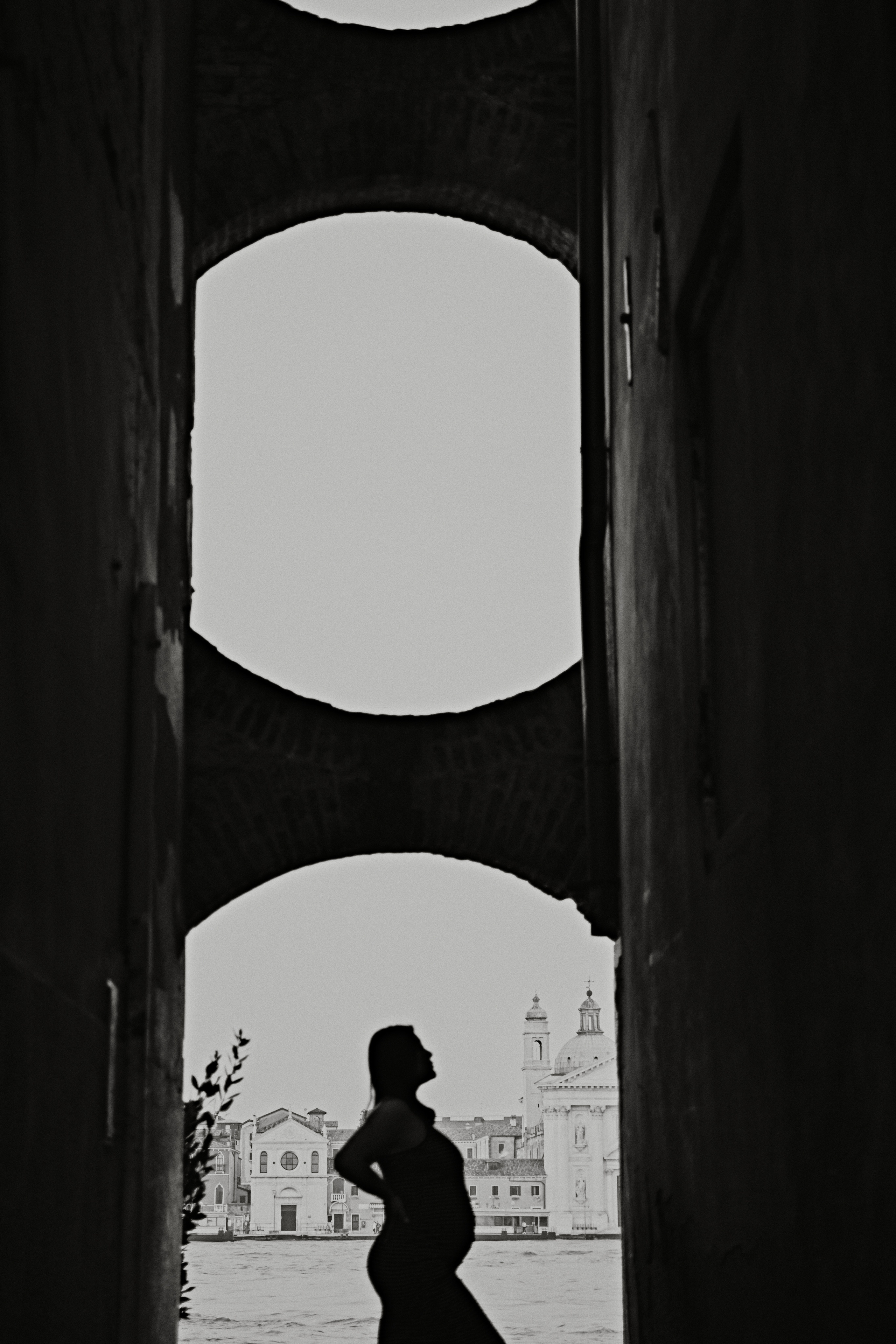 A radiant young woman in jeans stands amidst the picturesque Colonna area of Venice. Her hands gently cradle her growing belly, a symbol of the new life growing inside. The soft light of the sun filters through the narrow alleyways, casting a warm glow on her face. The calm waters of the canal reflect the colorful buildings and arch bridges, alluding to the timeless beauty and serenity of the city. The woman's peaceful expression captures the joy and anticipation of impending motherhood, a moment frozen in time amidst the romantic backdrop of Venice.