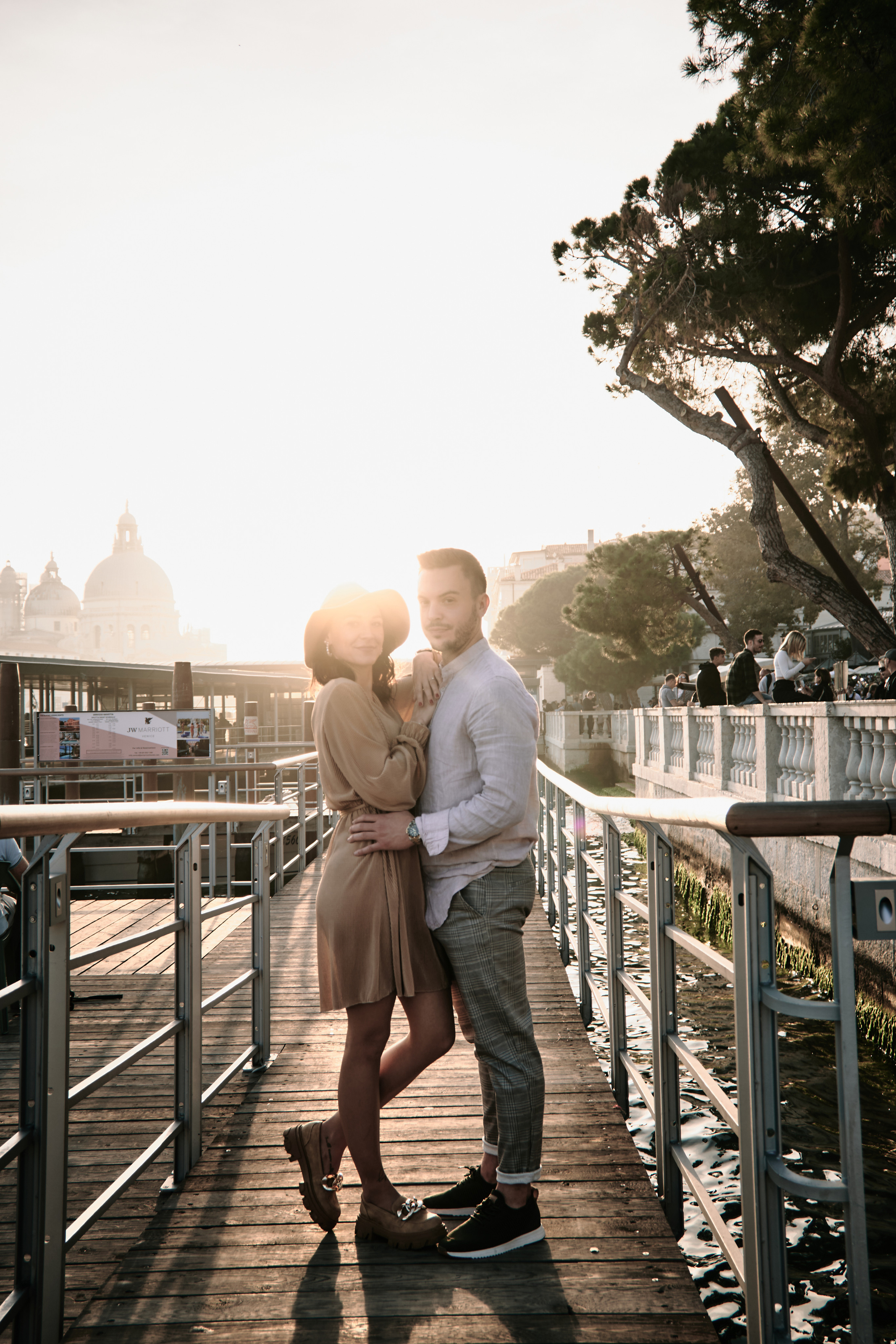 A couple stands on the edge of a beautiful lagoon, with the iconic Columns of San Marco and San Todaro in the background. The warm, golden light of the sun sets behind them, casting a romantic glow over the scene