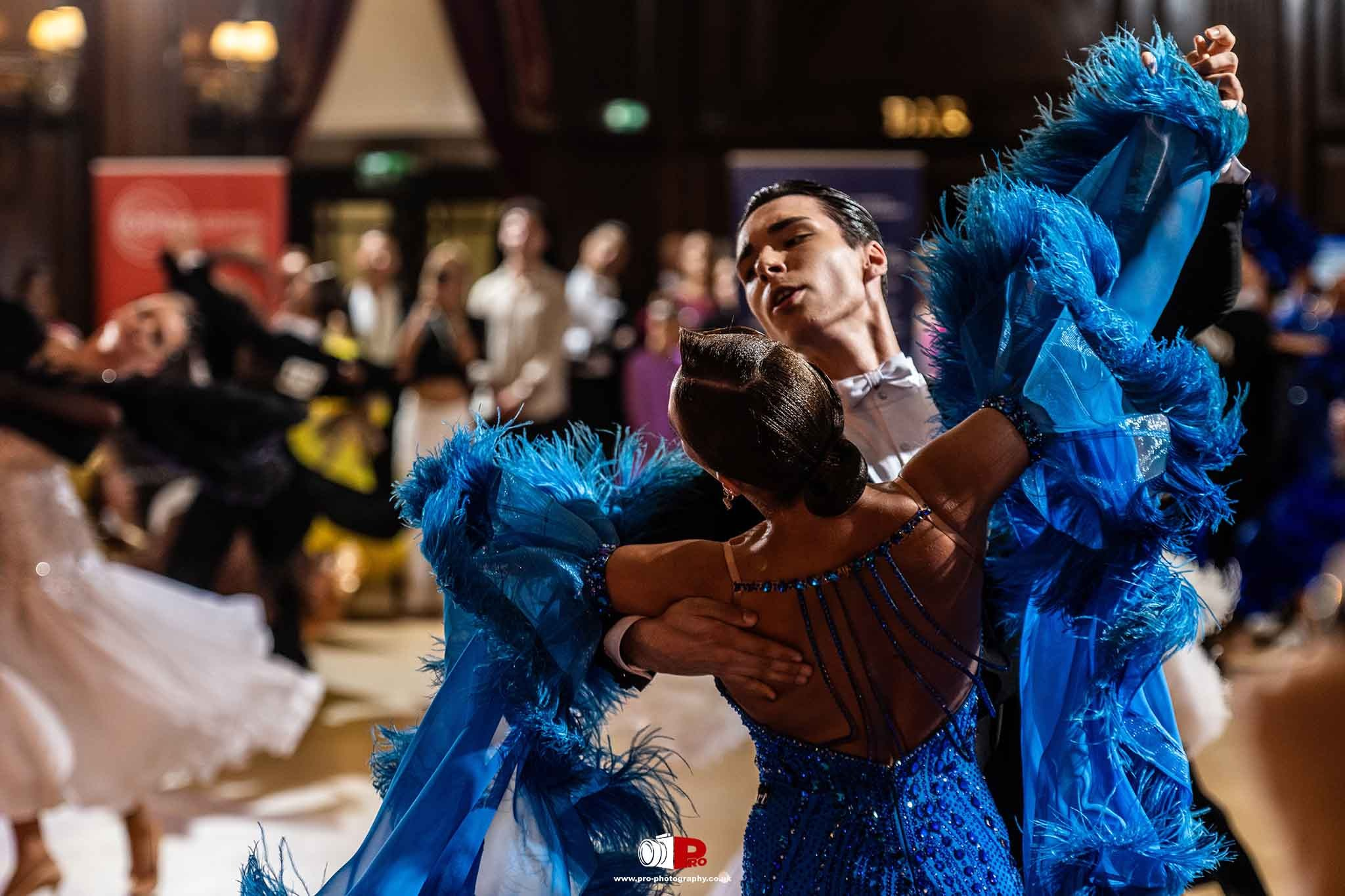 A ballroom couple gracefully performing in a rich blue feathered costume at a grand dance event.