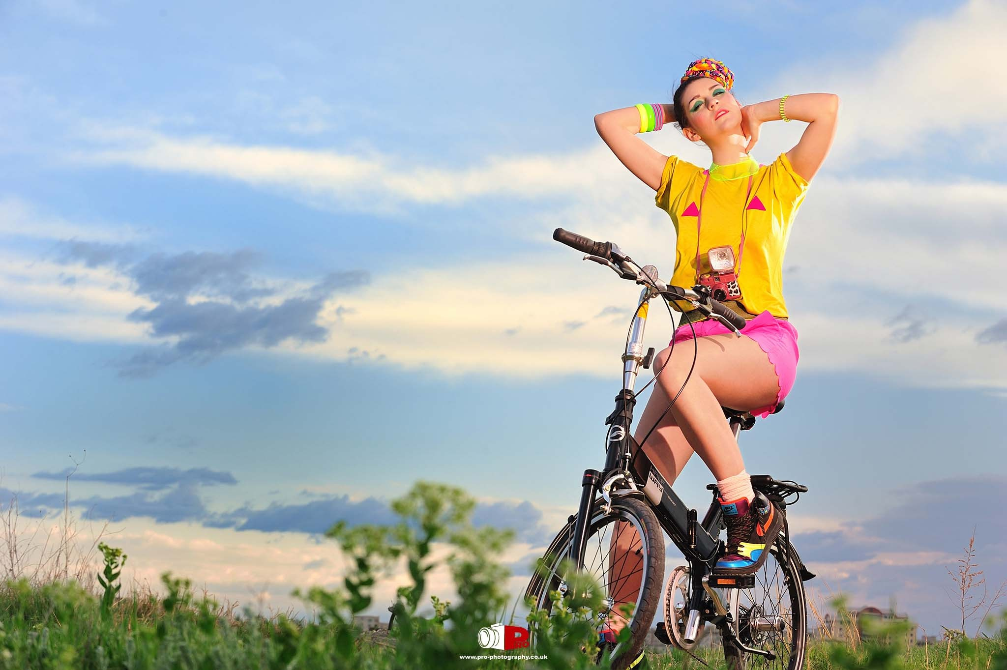 A cheerful young woman dressed in colourful attire, posing on a bicycle against a serene sky and green field backdrop.